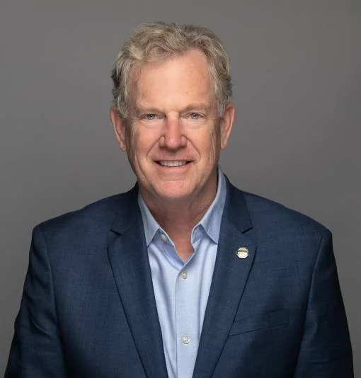 Headshot of a middle-aged man with gray hair, wearing a blue suit and a light blue dress shirt, smiling against a gray background.