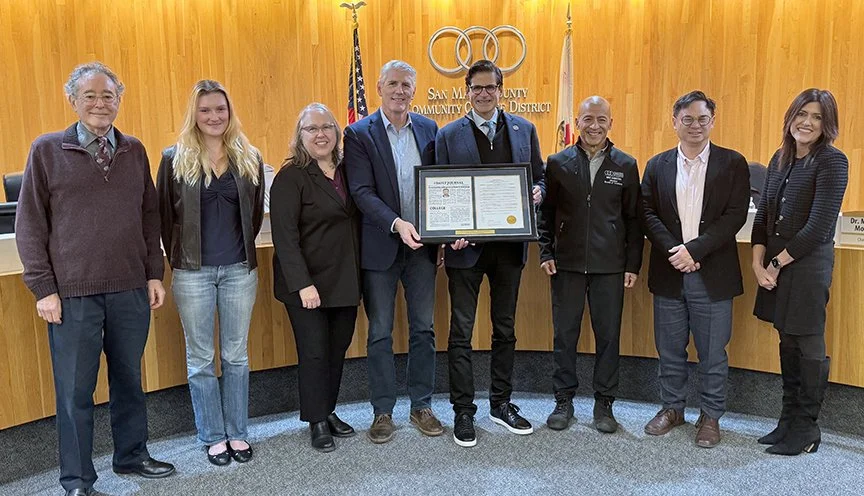 Group of eight people standing in a line in a wood-paneled room, with a man in the center holding a framed certificate or award. Behind them are the American flag, the California state flag, and the San Mateo Community College District emblem.
