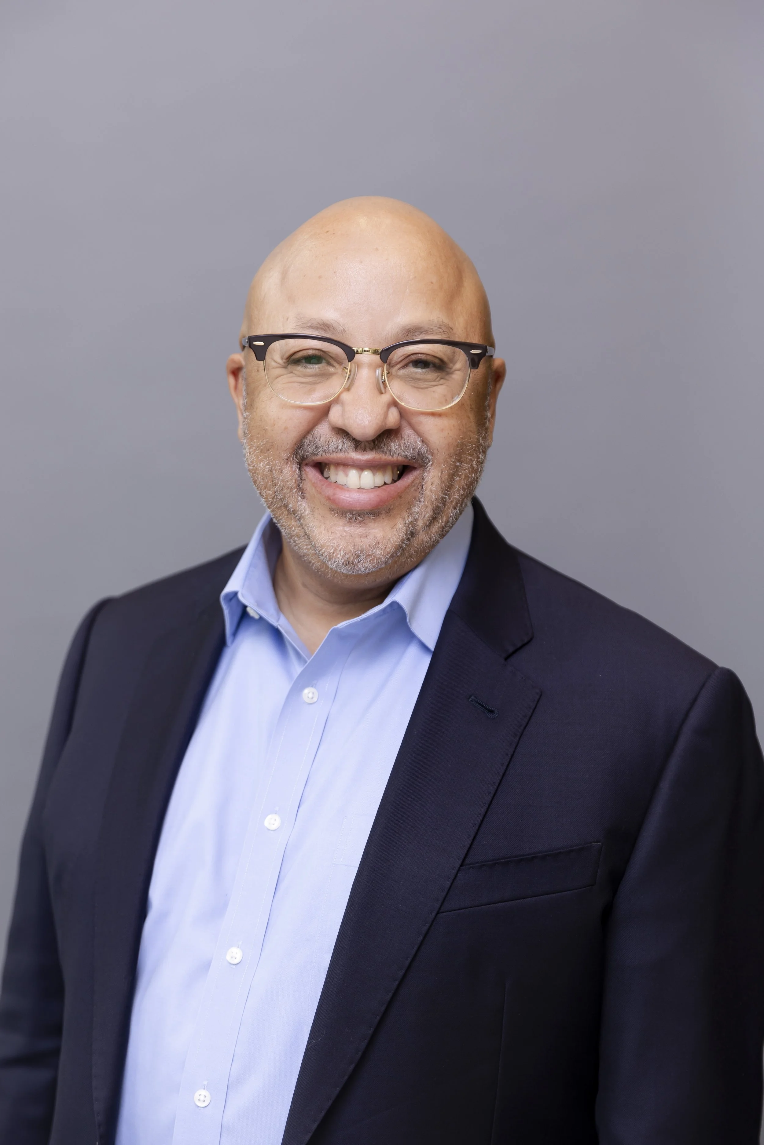 A smiling middle-aged man with a bald head, glasses, and a beard, dressed in a light blue dress shirt and a dark blazer, standing against a grey background.