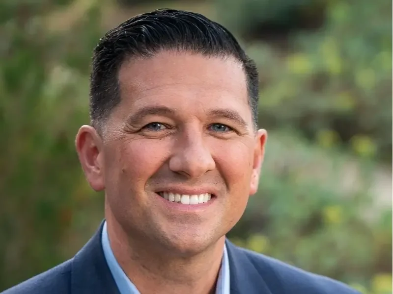 A smiling man with short dark hair, wearing a dark suit and light blue shirt, outdoors with blurred green and brown background.