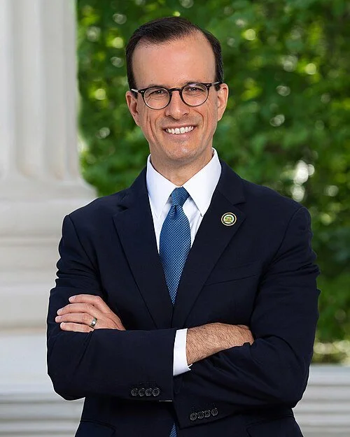A man in a navy suit and blue tie, wearing glasses, standing outdoors with crossed arms, smiling in front of greenery.