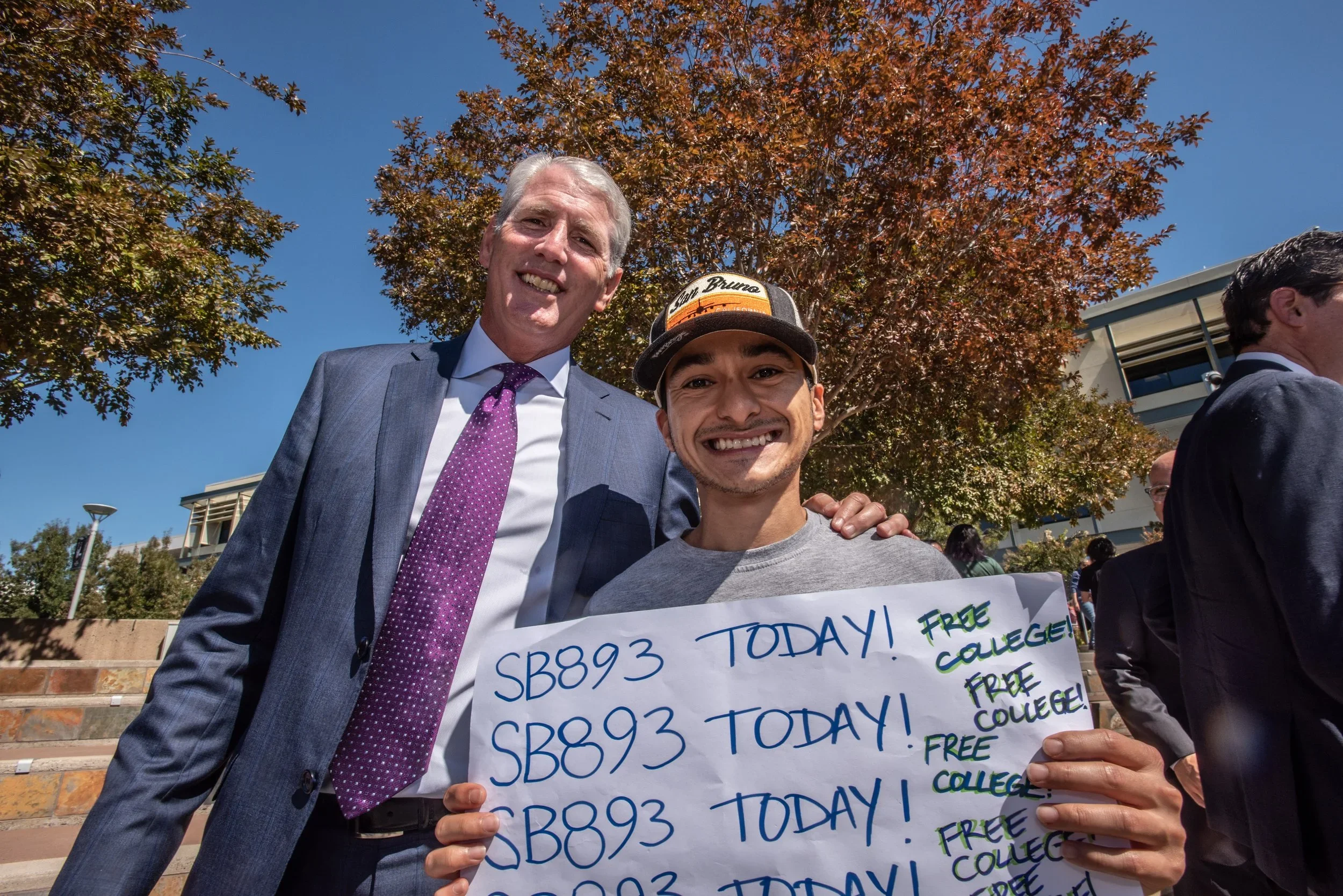 Two smiling men standing outdoors under a tree on a sunny day, one in a suit and the other holding a sign that reads 'SB893 TODAY! FREE COLLEGE!'. The man in a suit has gray hair and is wearing a purple tie, while the other is wearing a gray t-shirt and a baseball cap.