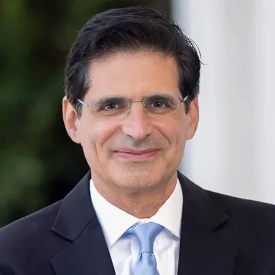 A man in a dark suit, white shirt, and light blue tie smiling, with glasses and dark hair, standing outdoors near a white building or column.