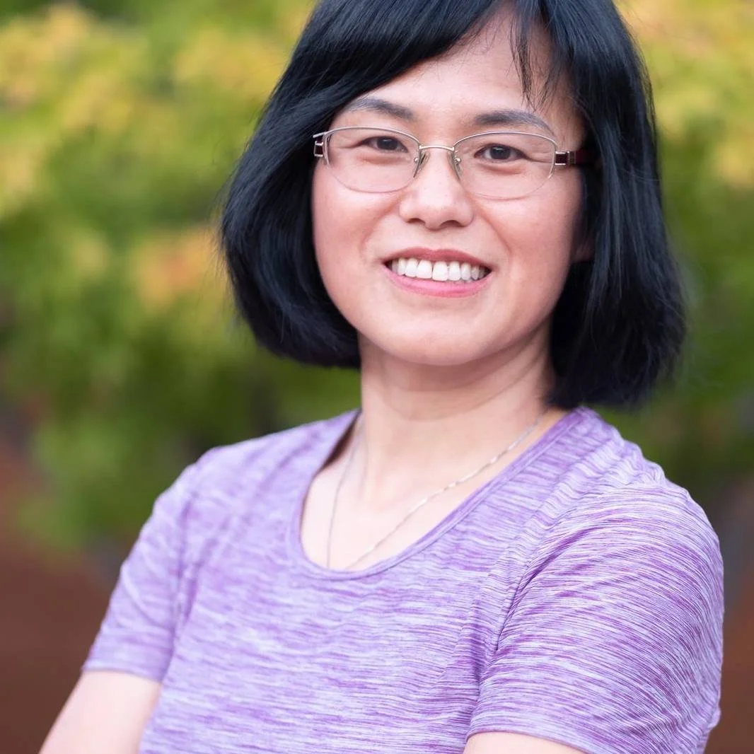 A woman with short black hair, wearing glasses and a purple shirt, smiling outdoors with blurred trees in the background.