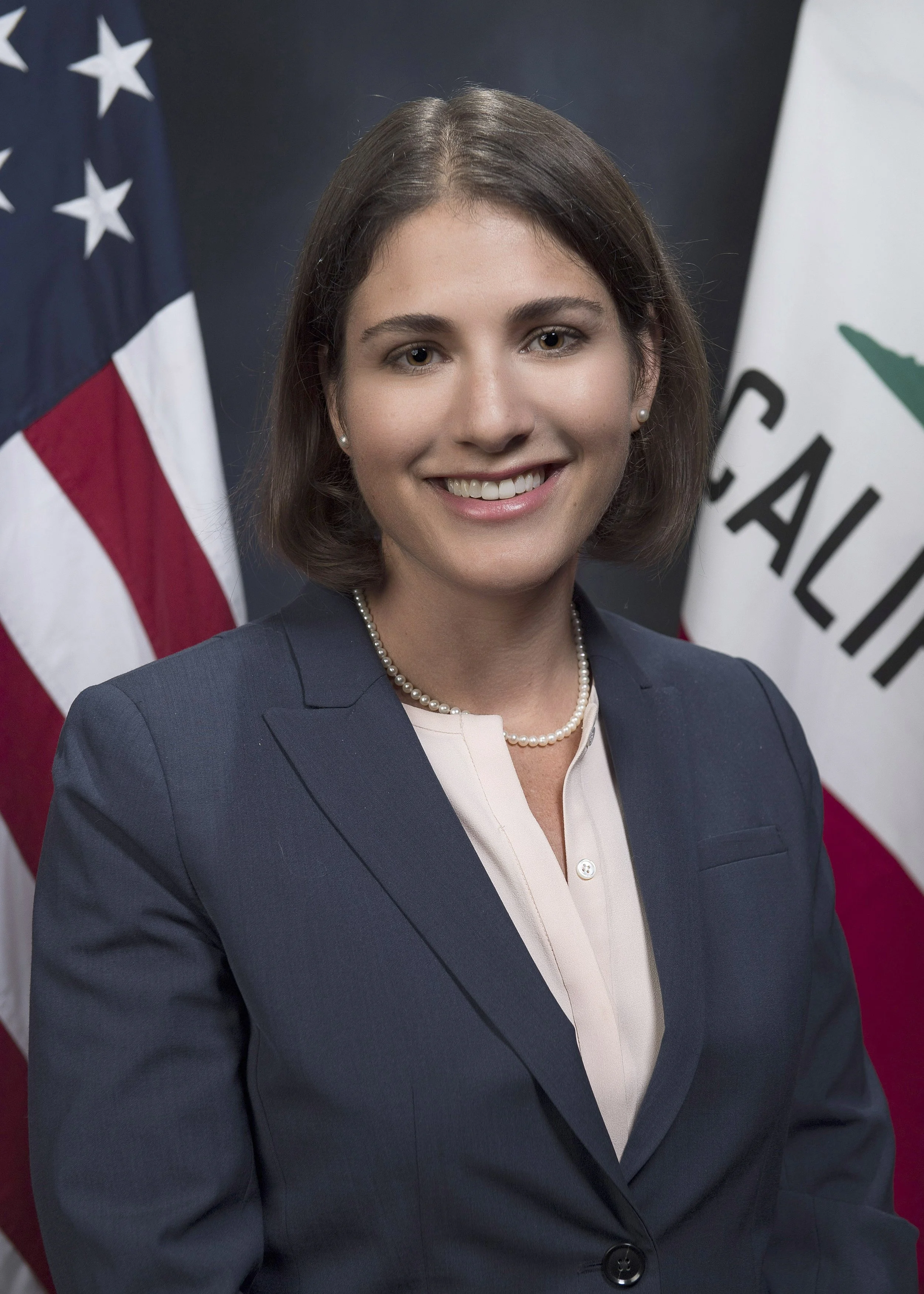 A smiling woman in a navy blazer and pearl necklace standing in front of American and California flags.