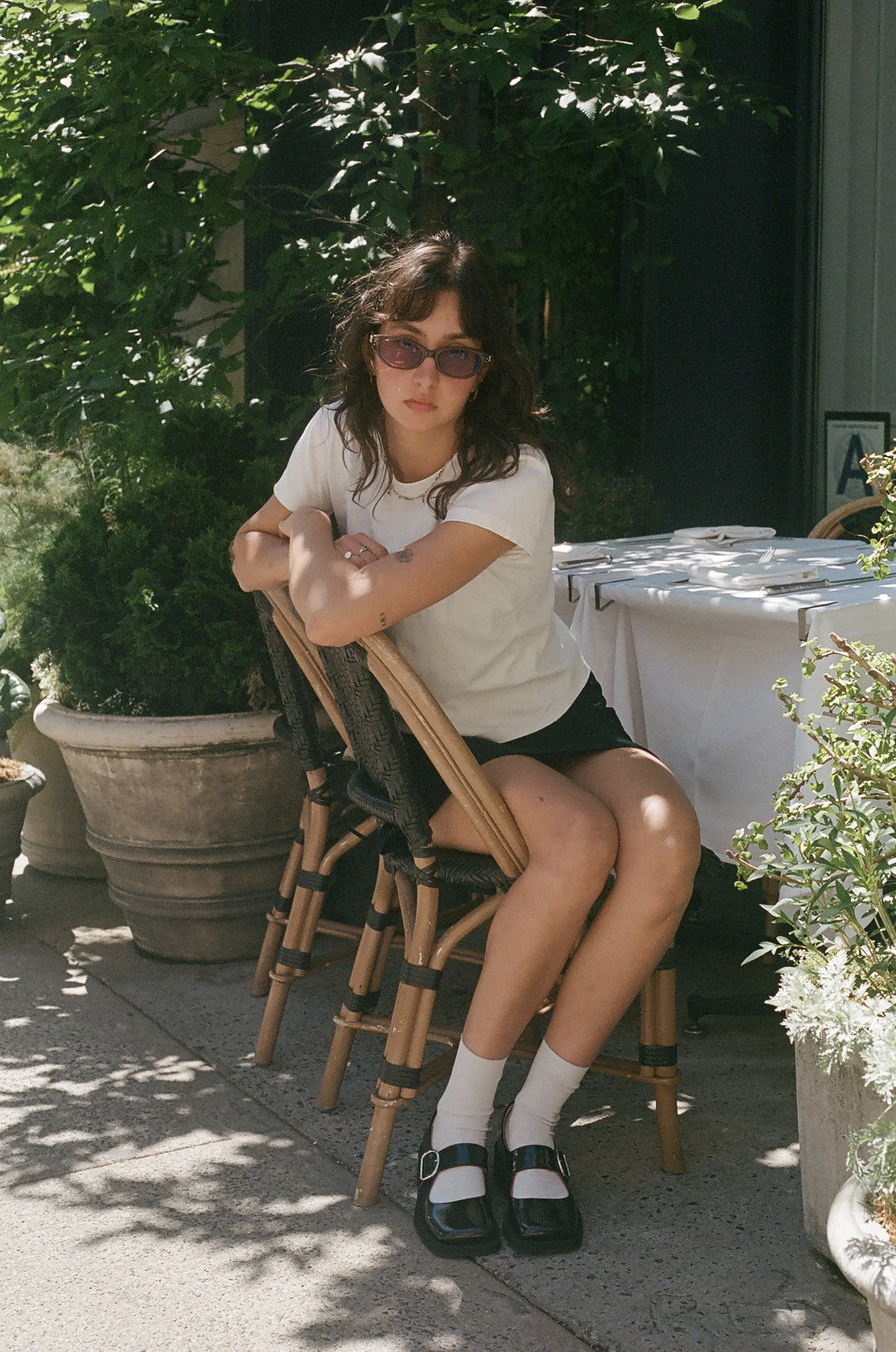 A woman with wavy dark hair wearing pink-tinted sunglasses, a white t-shirt, black shorts, white socks, and black platform shoes, sitting on a wicker and wood chair at an outdoor dining area surrounded by greenery, with a table set with plates, utensils, and glasses behind her.