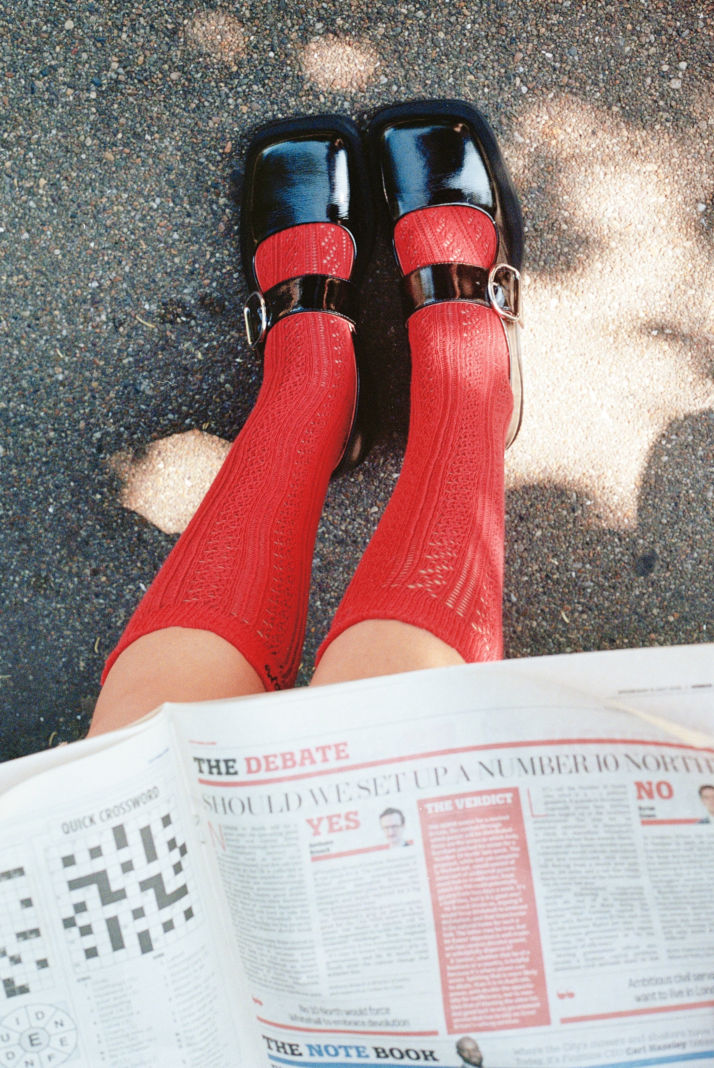 Person wearing red thigh-high socks and black patent leather shoes with metal buckles, reading a newspaper outdoors on a gravel surface.