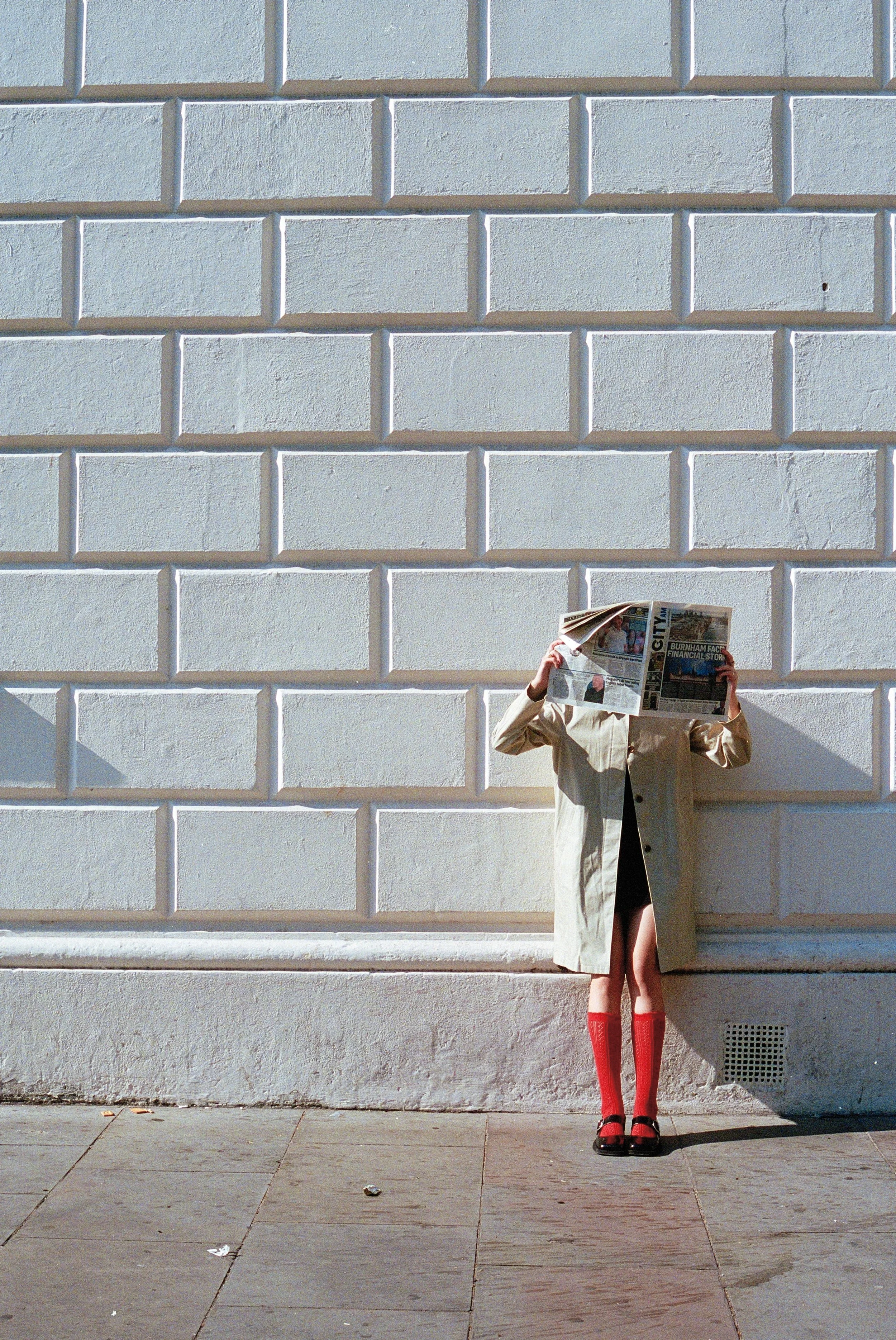 Person wearing a beige trench coat, red socks, and black shoes standing against a plain white brick wall, holding a newspaper overhead that obscures their face.