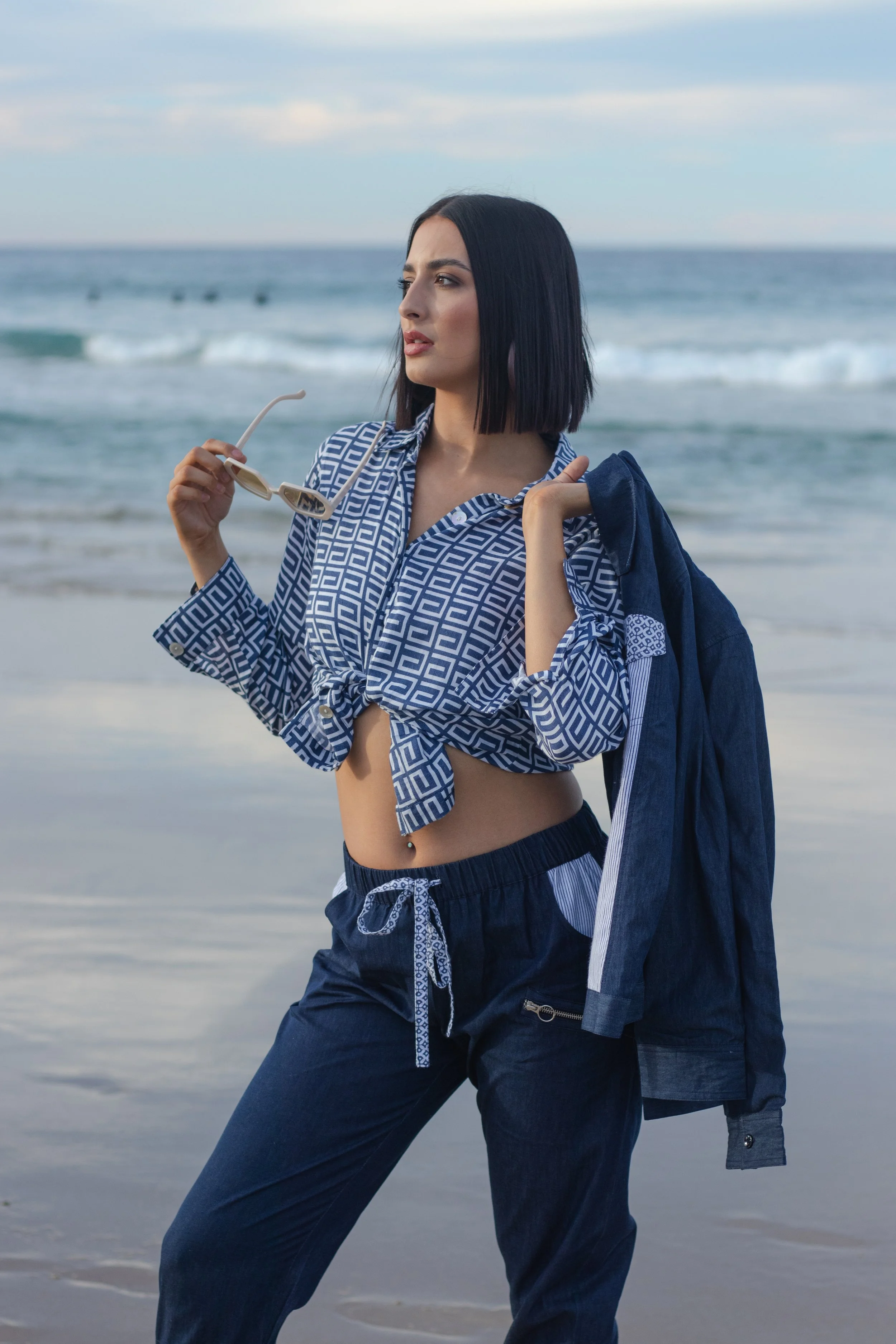 A woman with straight black hair, wearing a blue patterned button-up shirt tied at the waist and navy sweatpants, holds a pair of sunglasses while standing on a beach with ocean waves in the background.