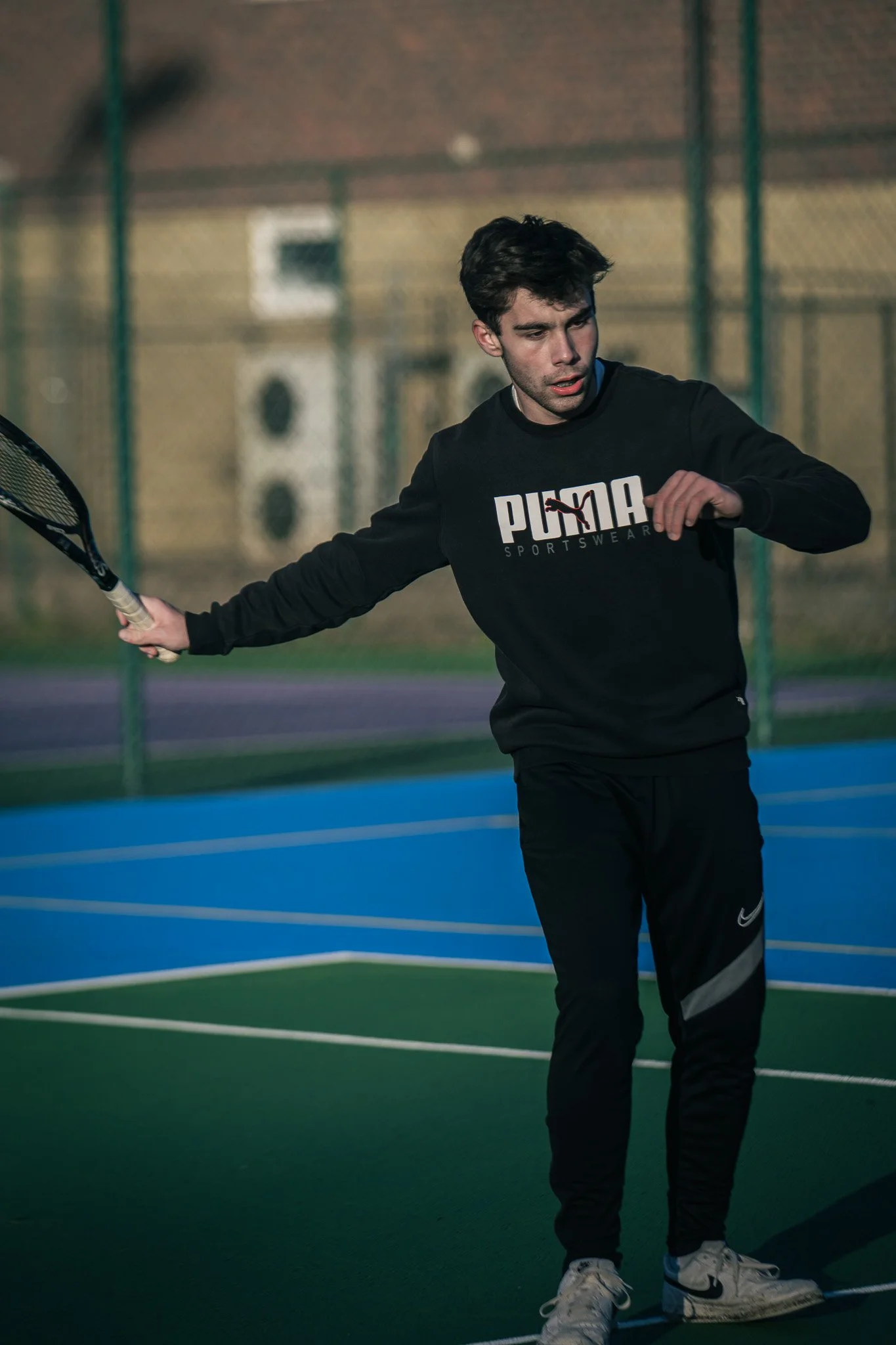 A young man playing tennis on an outdoor court during evening or early morning, wearing a black Puma sweatshirt and black Nike pants, holding a tennis racket in his right hand, preparing to hit the ball.