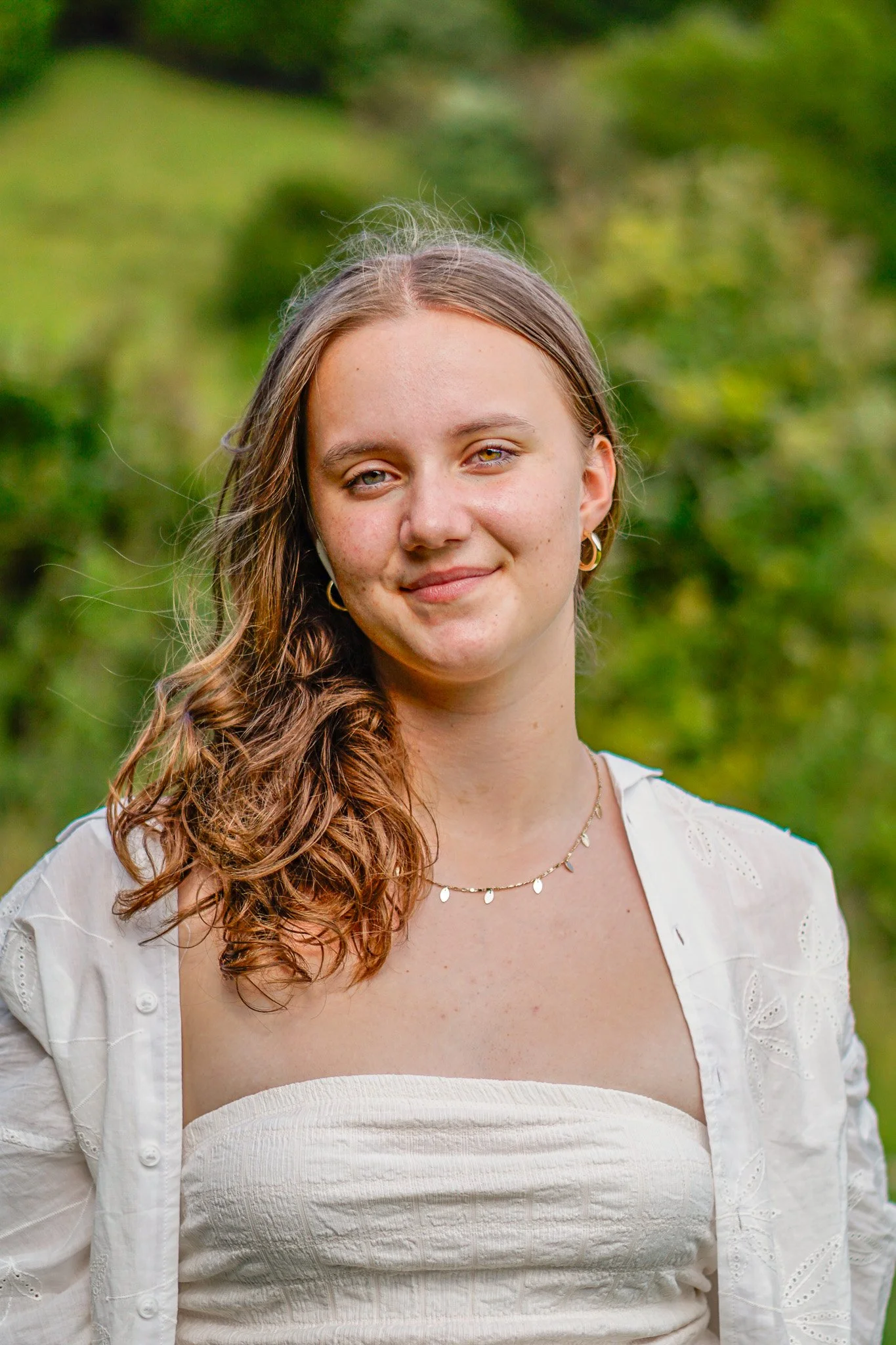 A young woman with long, curly brown hair and light skin, smiling gently, standing outdoors with a blurred green background.