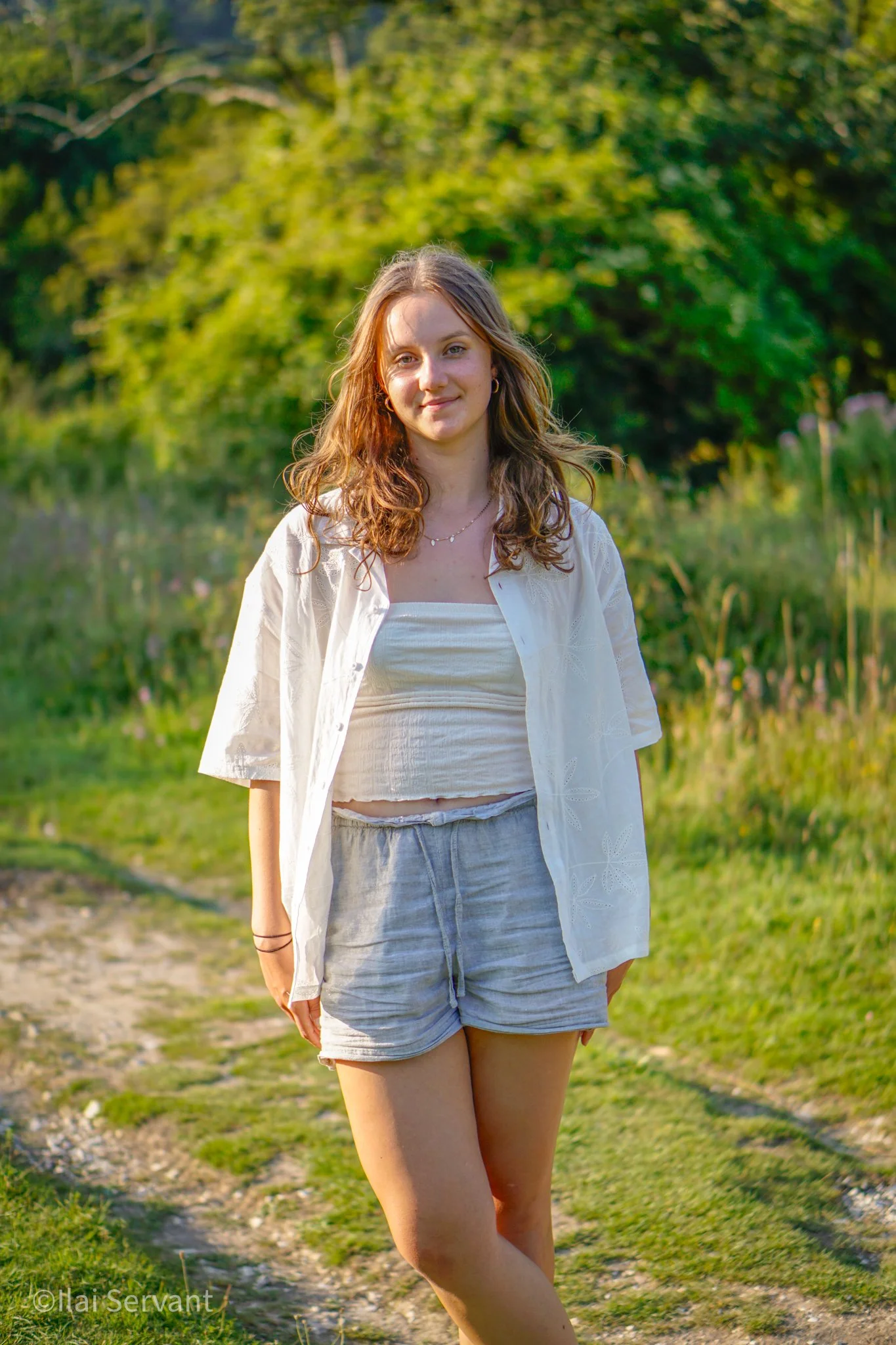 A young woman with curly brown hair wearing a white button-up shirt open over a cream crop top, gray shorts, standing outdoors on a grassy path with green trees in the background during the daytime.