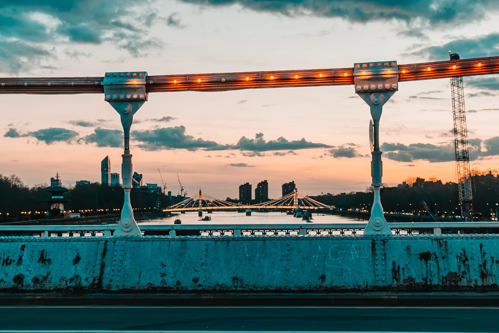 City skyline viewed from a bridge during sunset with a river, modern buildings, and a distinctive cable-stayed bridge in the background.