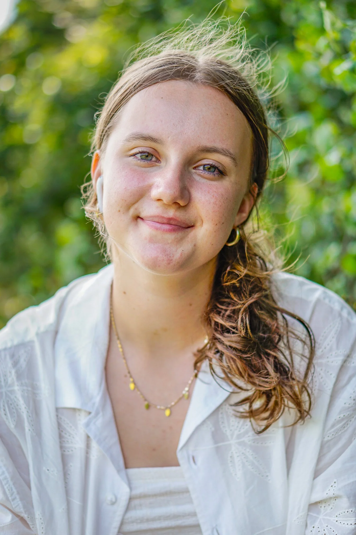 A young woman with light skin and wavy light brown hair, smiling outdoors with green foliage in the background. She wears a white embroidered top and gold jewelry.