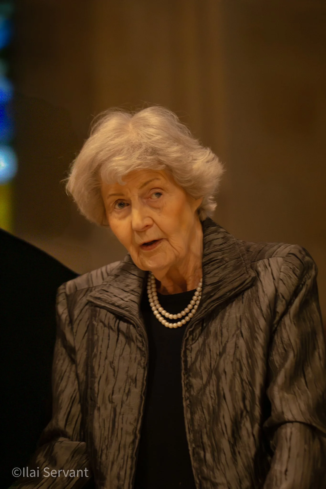 An elderly woman with gray hair, wearing a black top, a patterned gray jacket, and a pearl necklace, looks to the side with a thoughtful expression.