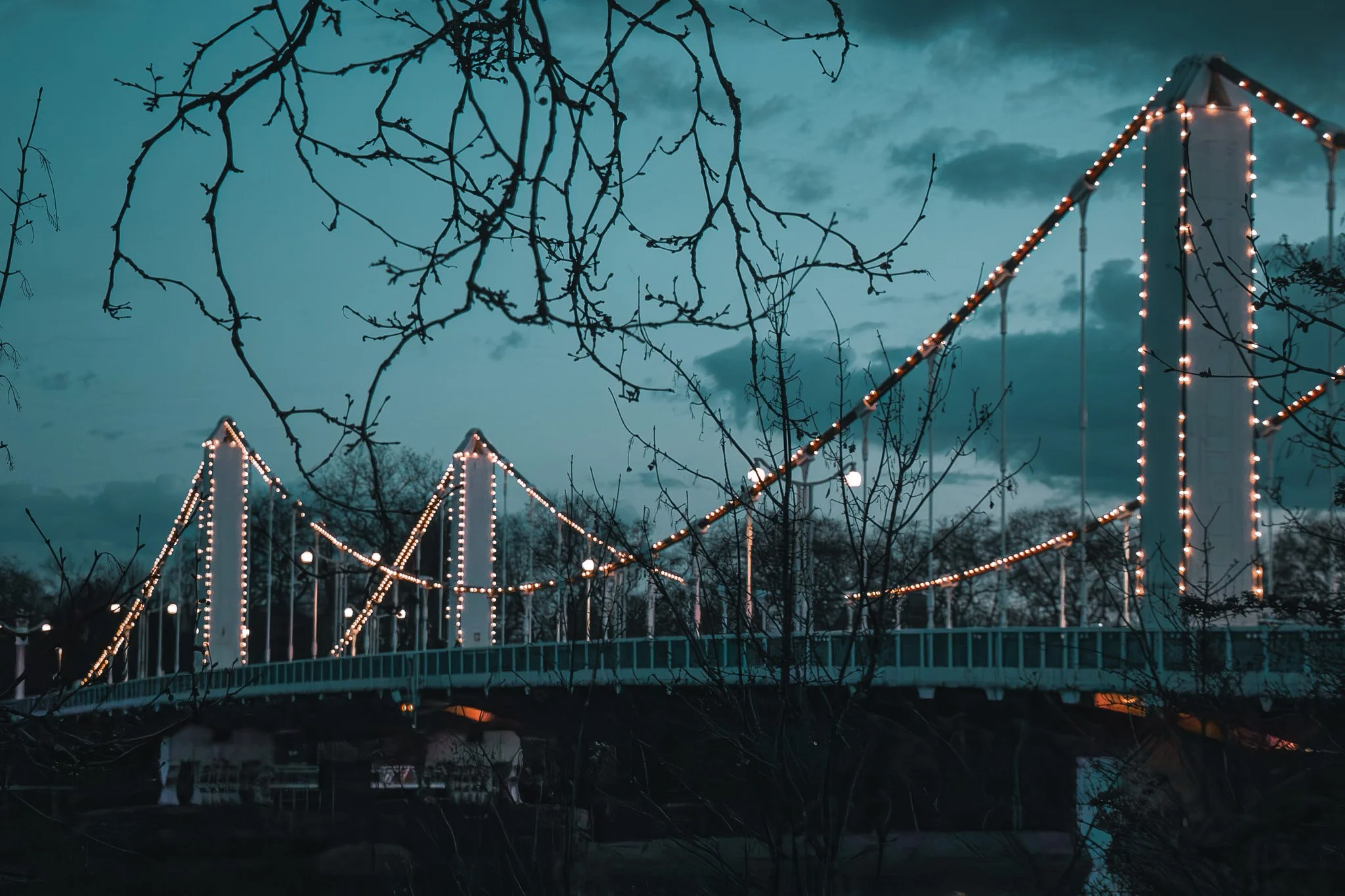 A suspension bridge decorated with string lights at dusk, with silhouetted trees and cloudy sky in the background.