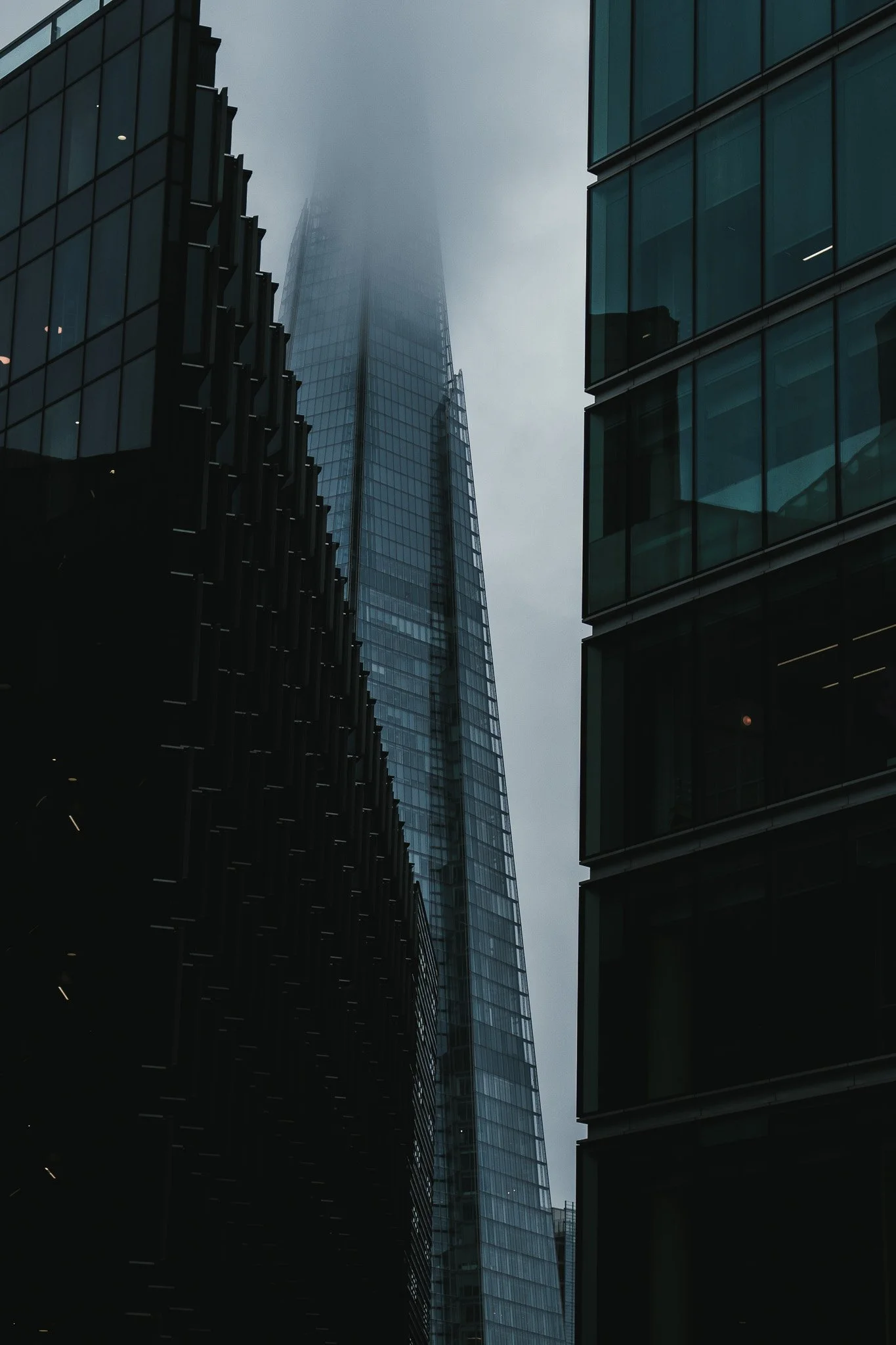 Tall modern skyscraper partially obscured by fog, viewed between other glass buildings in a city.