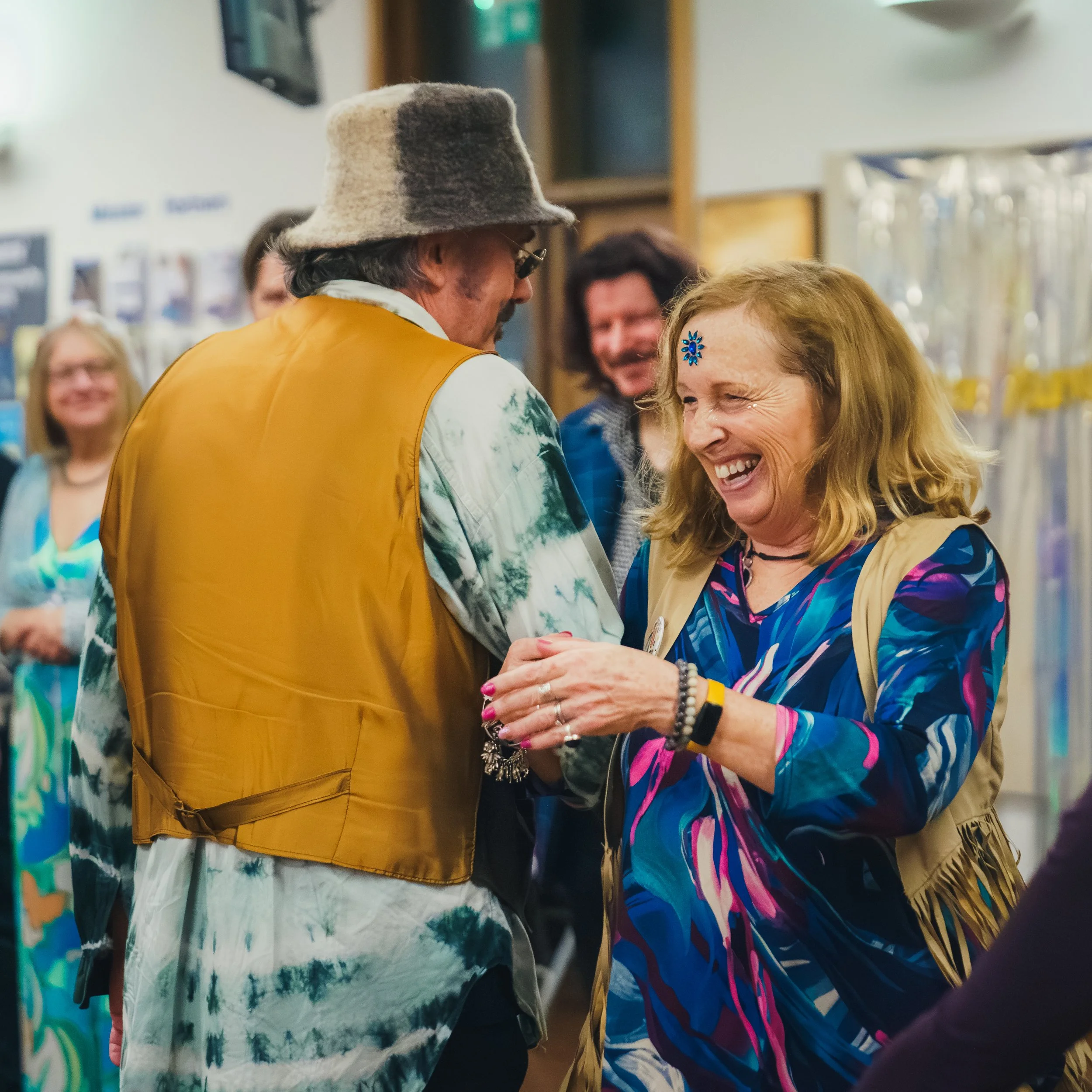 A group of people smiling and engaging in conversation indoors, with colorful clothing and accessories, including a woman with a blue and purple patterned jacket and a woman with a blue flower hair clip.