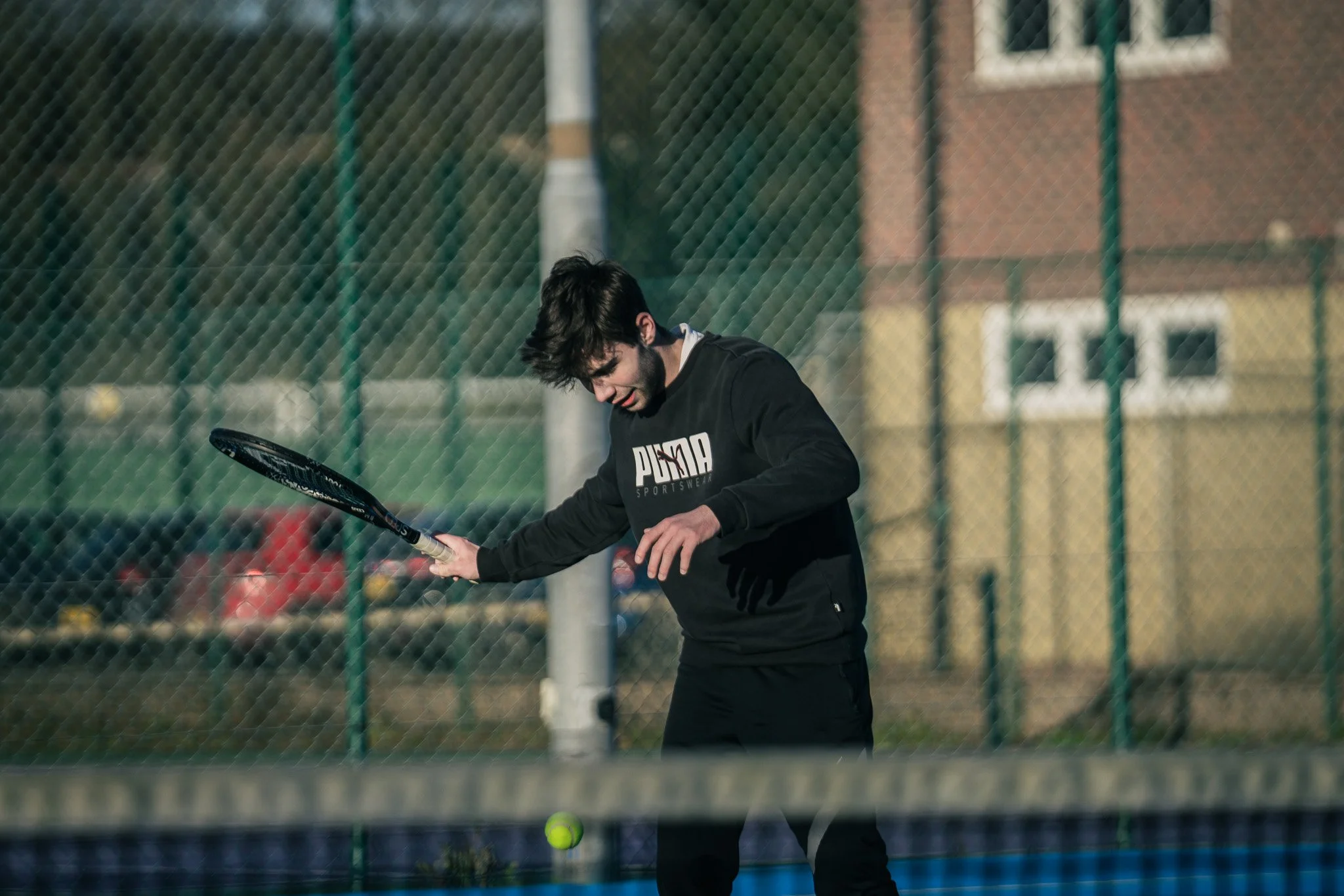 A young man is playing tennis outdoors on a court surrounded by a chain-link fence. He is dressed in a black Puma sweatshirt and black pants, holding a tennis racket, ready to hit a tennis ball near the net.