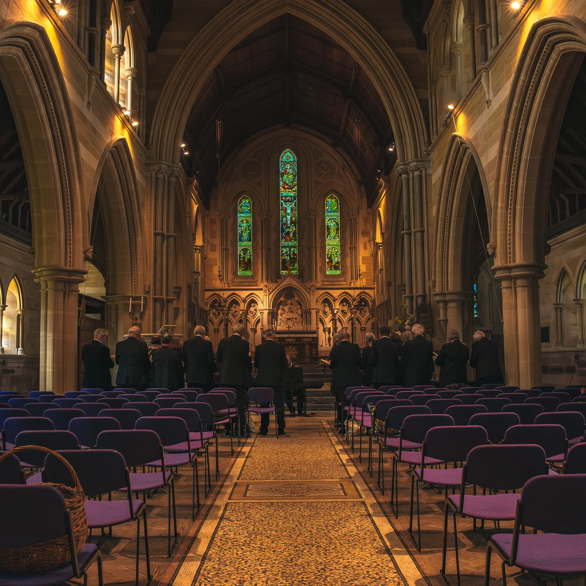 A group of people dressed in formal attire standing in a church with stained glass windows and high vaulted ceilings, facing the altar.