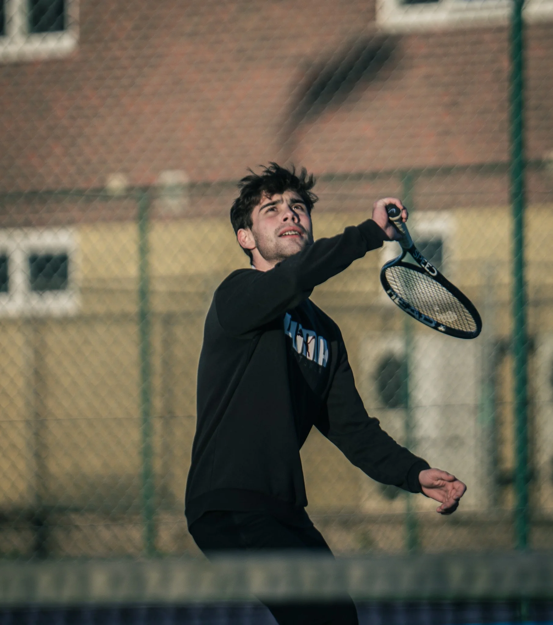 A young man playing tennis outdoors on a court at dusk, holding a tennis racket and preparing to hit the ball.