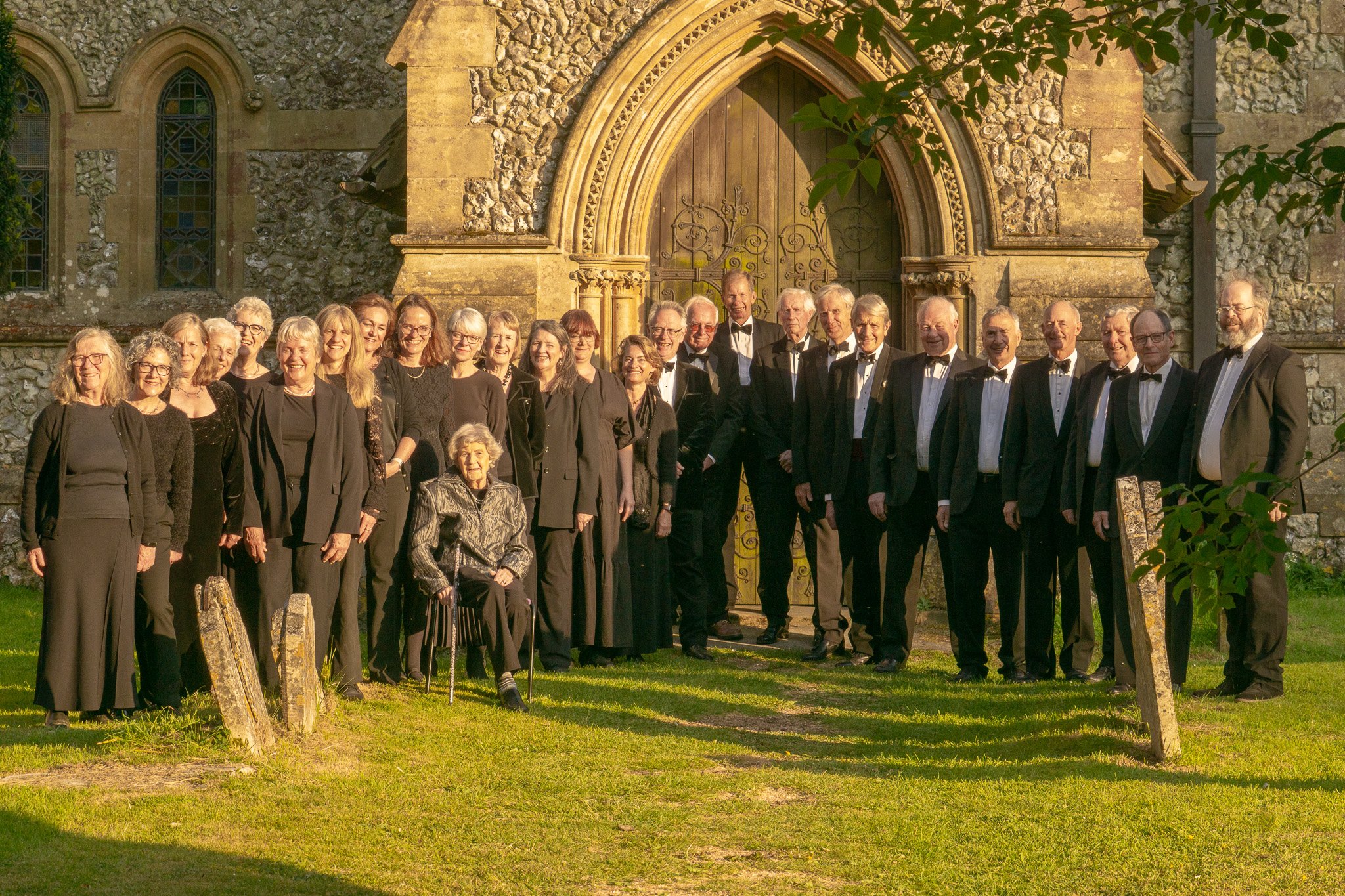 Group of older men and women in formal black attire, standing and sitting in front of a stone church with arched wooden door, during sunset.