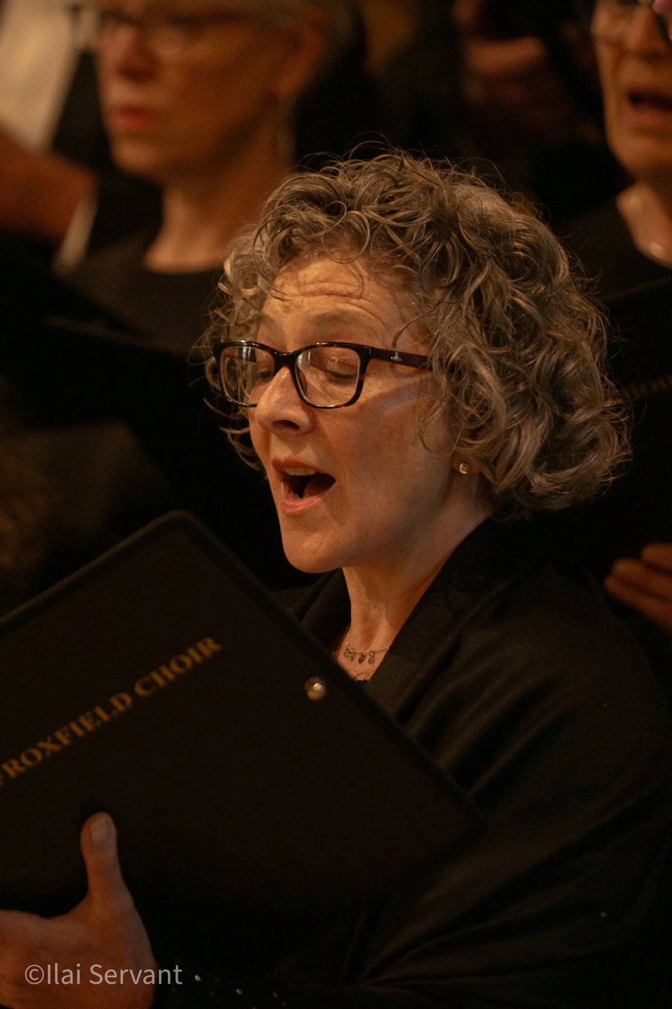 A woman with curly gray hair wearing glasses, singing from a black choir folder labeled 'ROXFIELD CHOIR' during a choir performance.