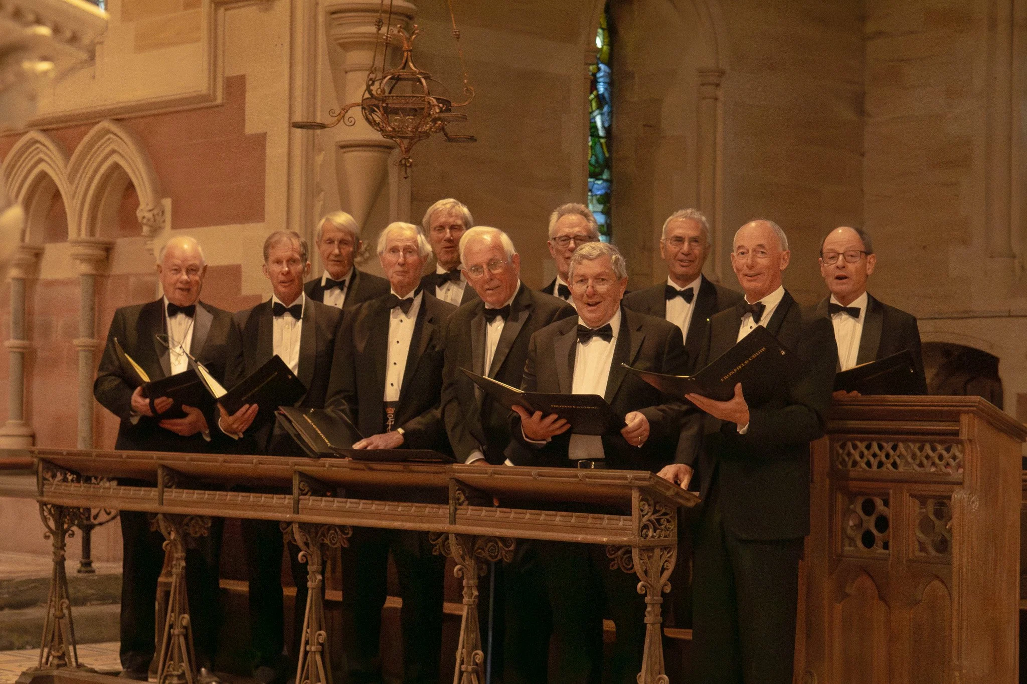 A group of elderly men in formal black tuxedos and bow ties singing together in a church choir, holding black folders or sheet music, standing behind a wooden choir stand with an ornate metal railing, inside a church with stained glass windows and go