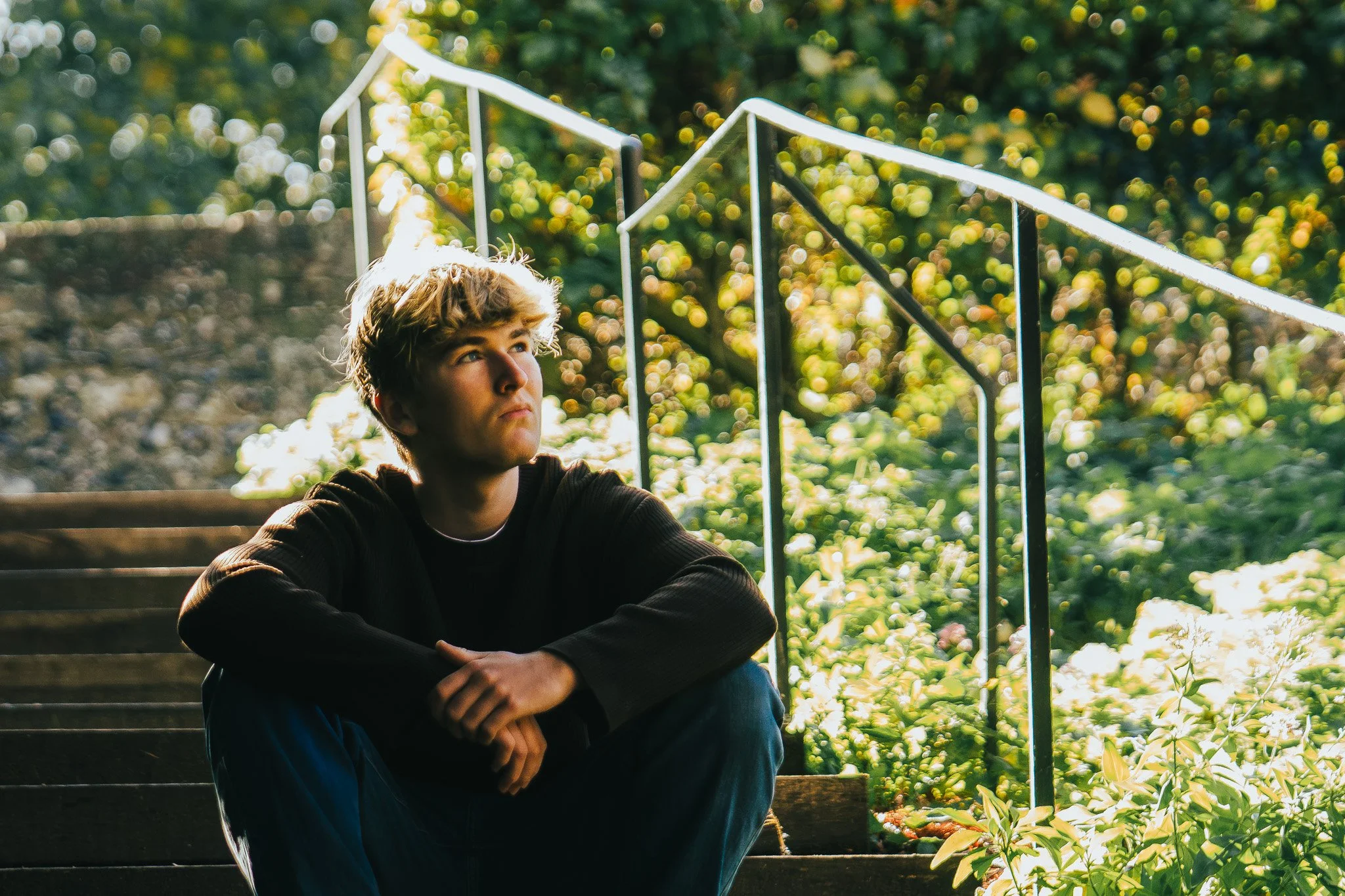 A young man with light brown hair sitting on outdoor stairs with greenery in the background, illuminated by sunlight.