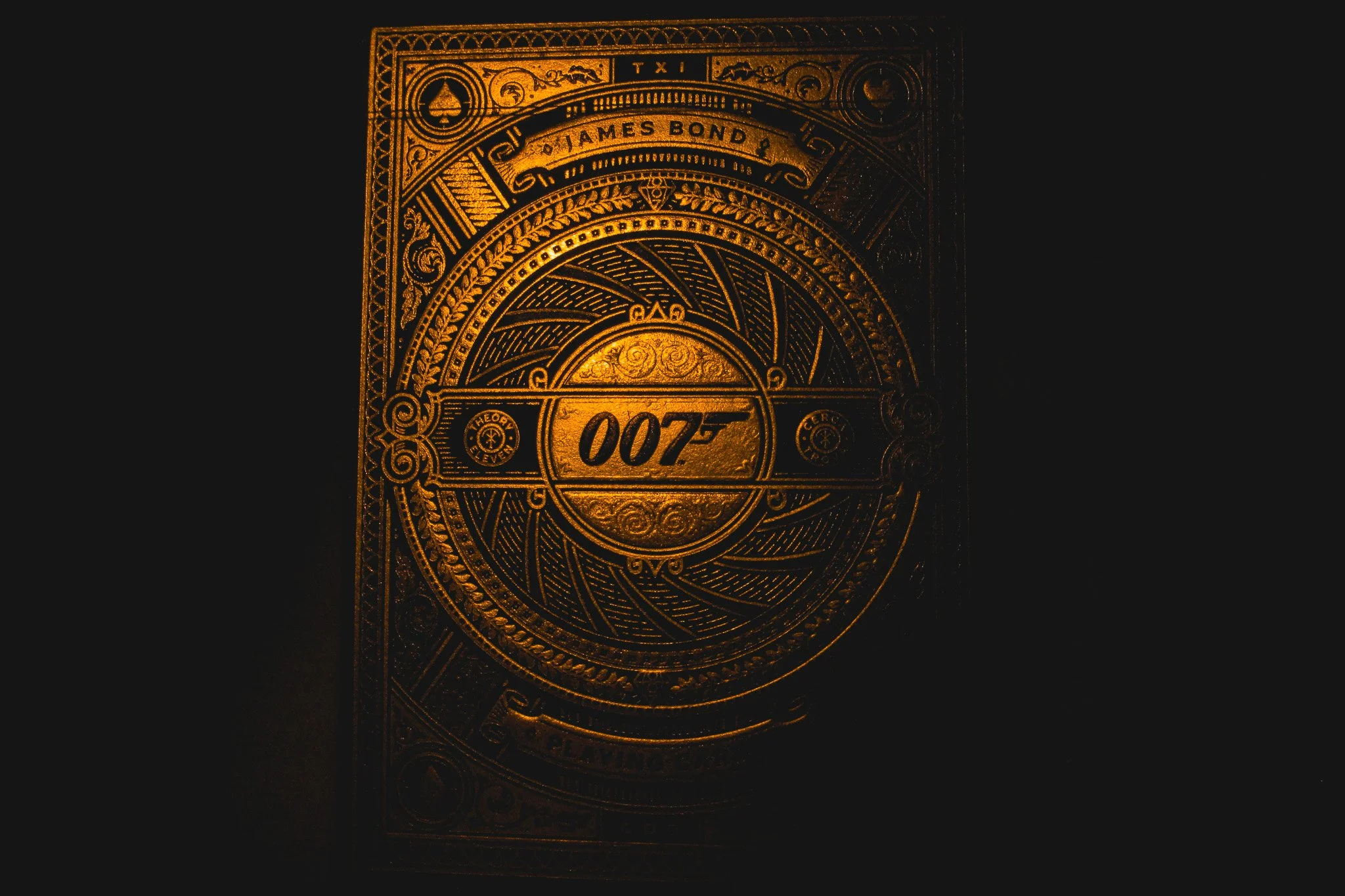 Close-up of a gold embossed design featuring James Bond's classic 007 logo with ornate patterns and the words "Theory Eleven" and "Cero" on a black background.