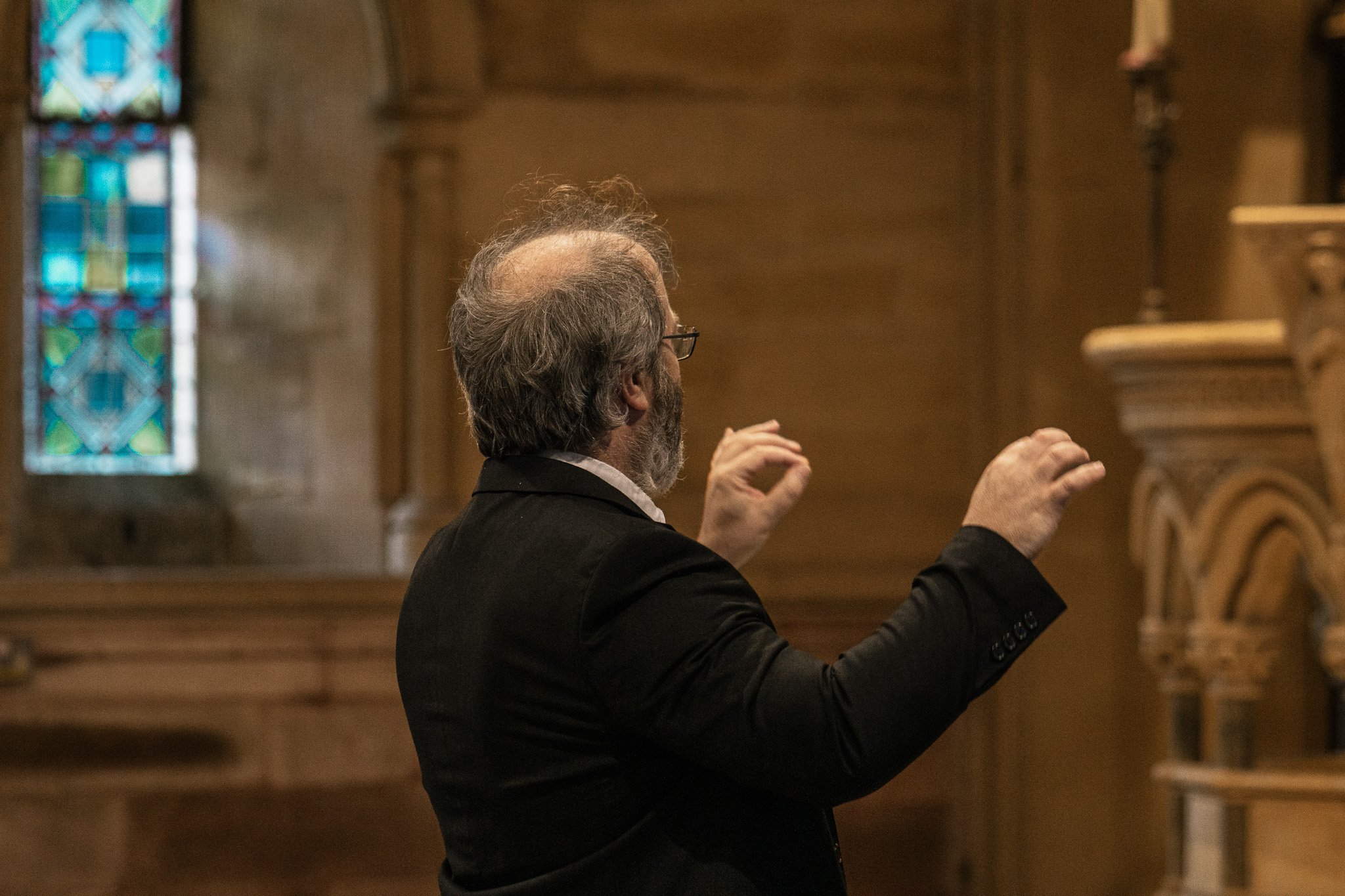 A man with gray hair and beard, wearing glasses and a black suit, is conducting or speaking inside a church or chapel with wooden walls and stained glass windows.