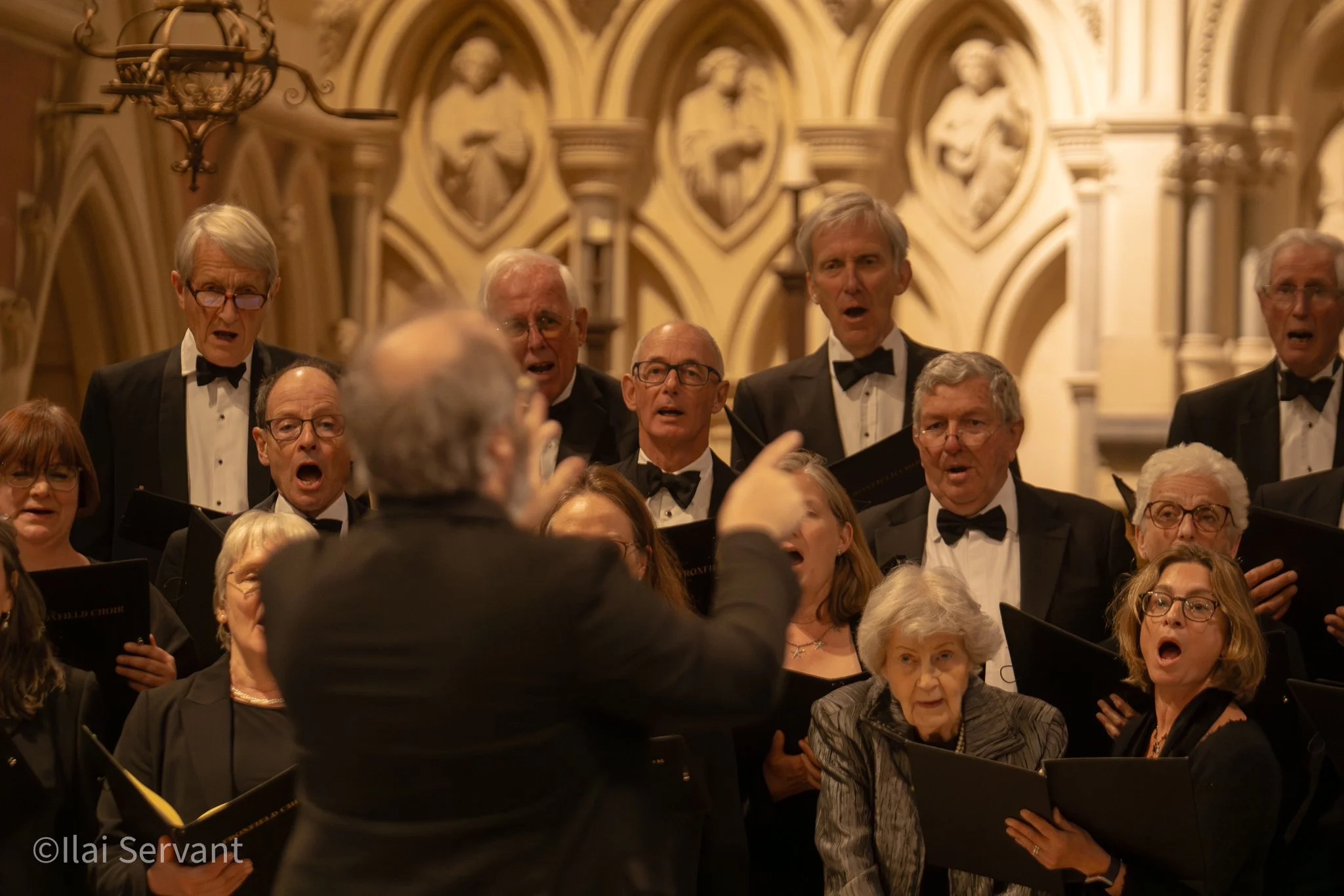 A choir performing in a church, led by a conductor. The choir members are wearing formal black attire, some with bow ties, and are singing while holding sheet music. The background features ornate wall decorations and sculptures.