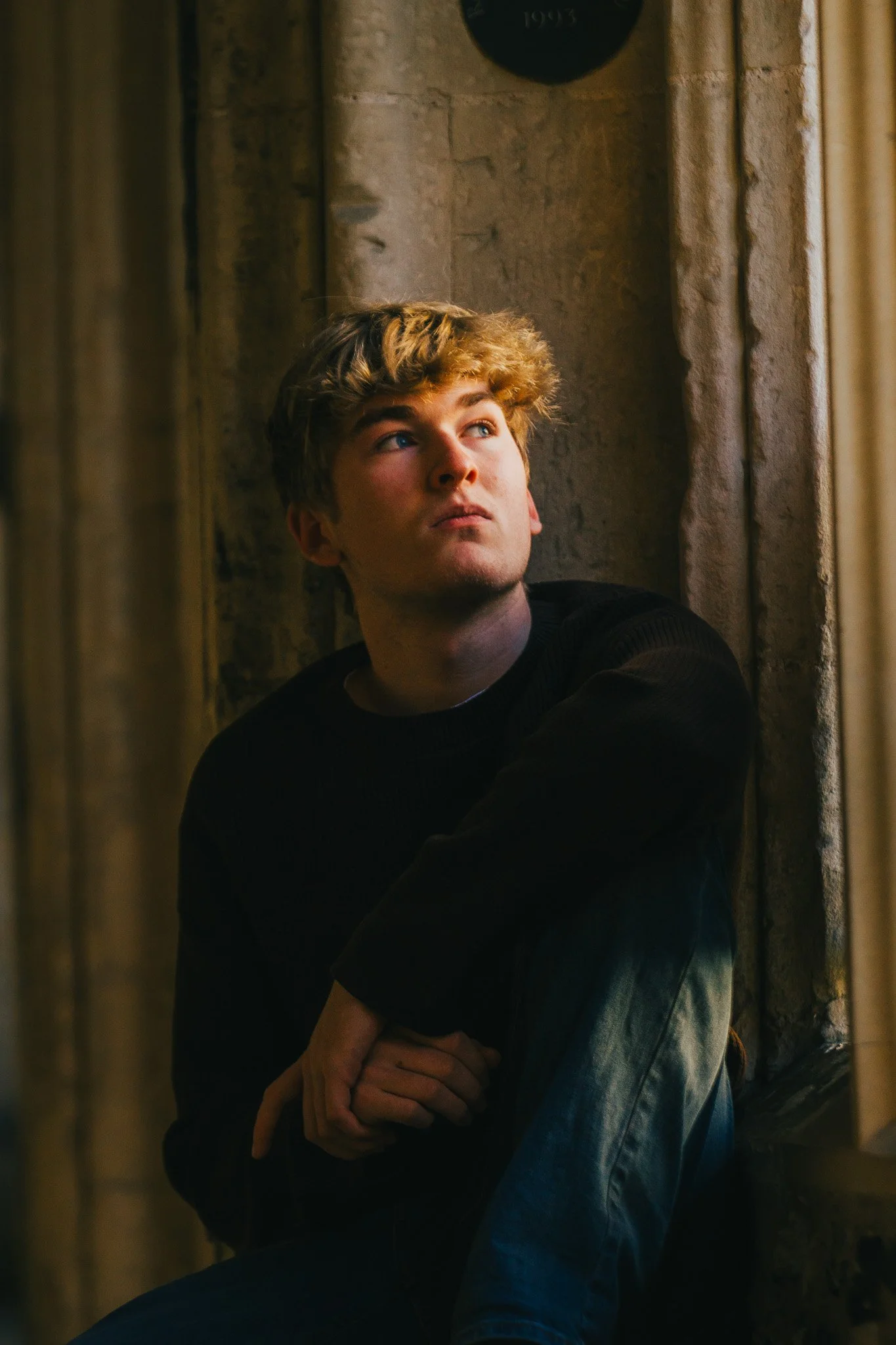 A young man with curly blonde hair sitting by a window, looking upward with a thoughtful expression.