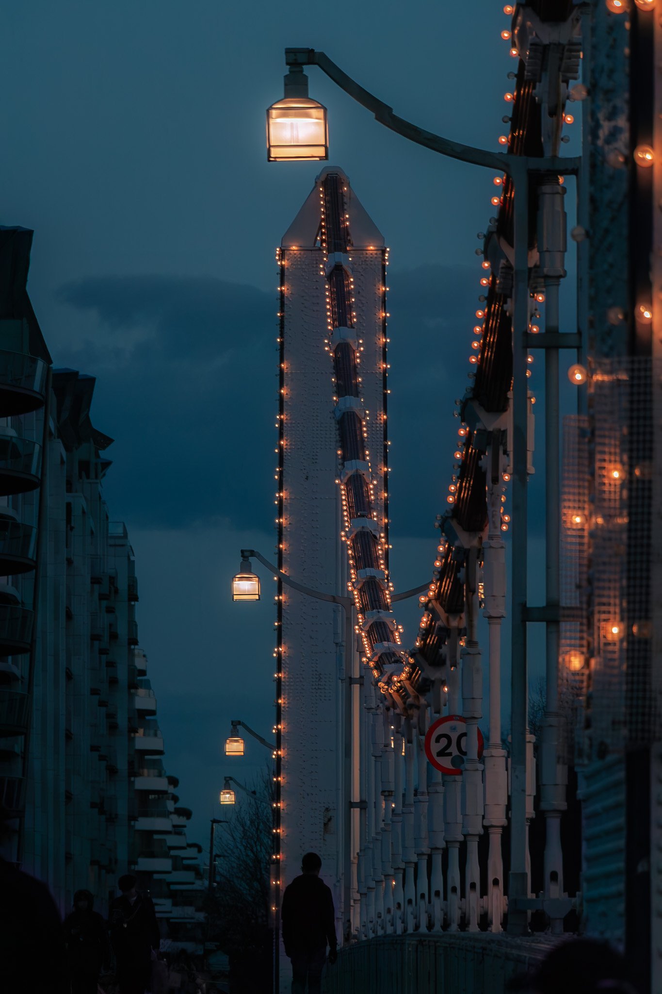 City street during evening with illuminated lamps and a tall structure decorated with lights in the background, and people walking along the sidewalk.