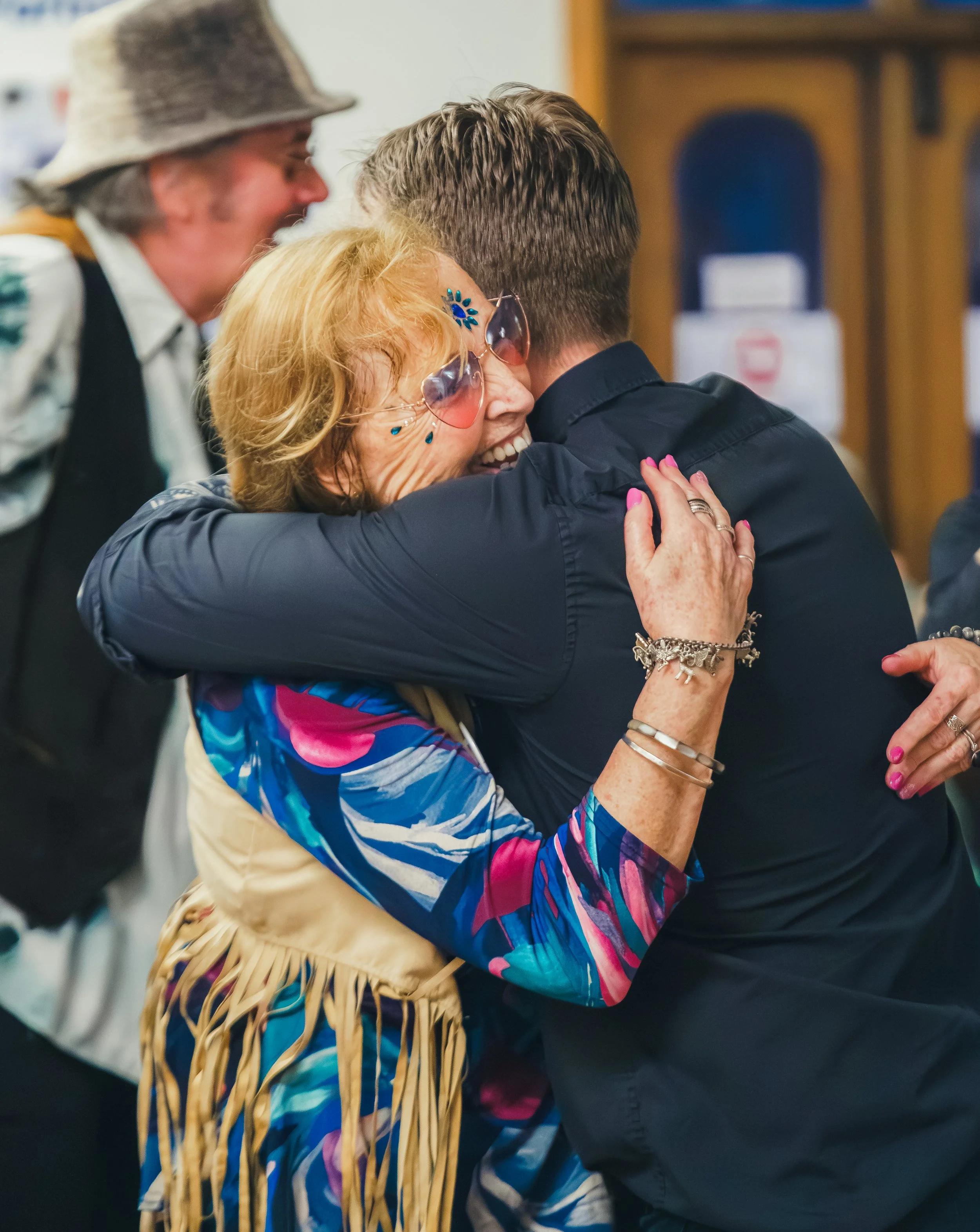 An elderly woman with colorful glasses smiling and hugging a young man, both appearing happy, while a man in a hat smiles in the background.