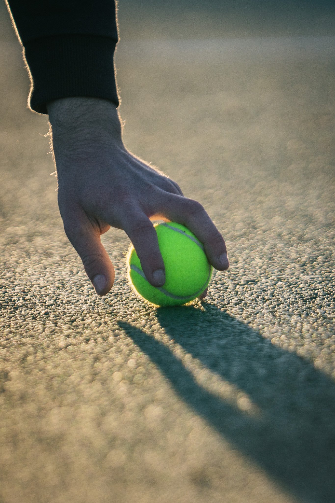 Close-up of a person's hand about to serve a tennis ball on a tennis court during sunset.