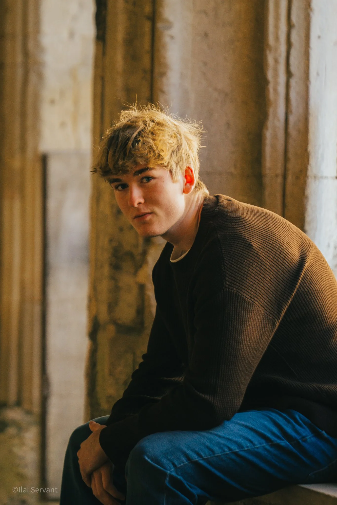 A young man with curly, blonde hair sits near a stone wall, looking directly at the camera with a neutral expression.