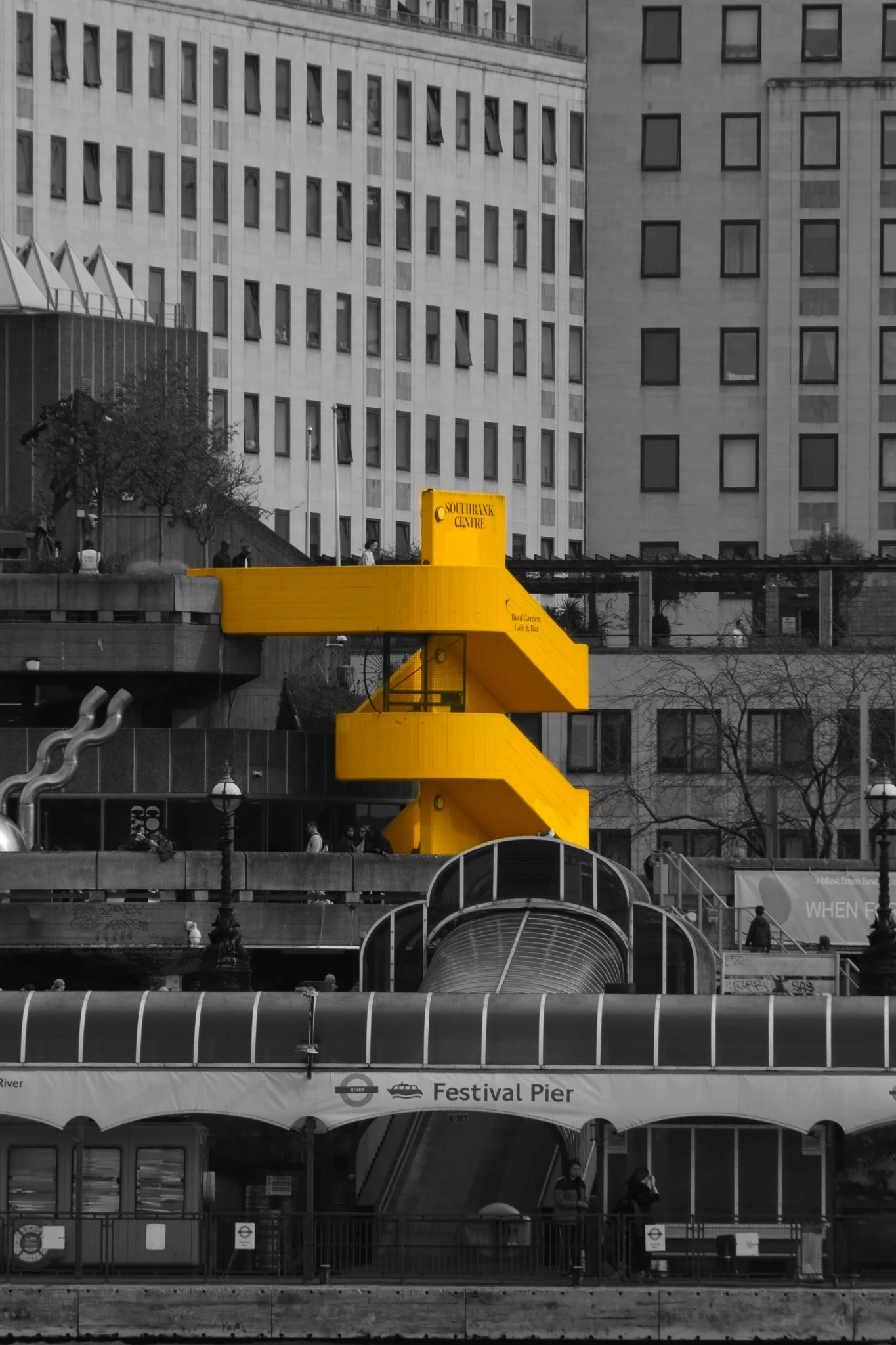 A black-and-white photo of a cityscape with tall buildings and a bright yellow staircase structure. The staircase is labeled 'Southbank Centre' and has multiple landings and spiral stairs. In the foreground, there's an entrance to a pier labeled 'Fes