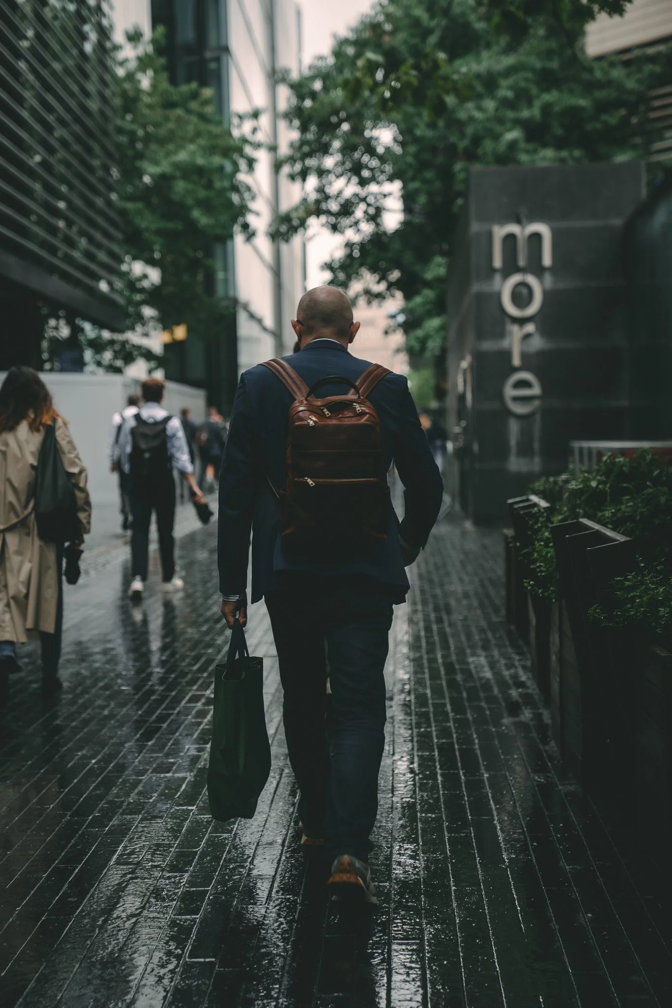 A man in a dark suit with a brown backpack walking on a wet city sidewalk, with other pedestrians and a building with signs visible.