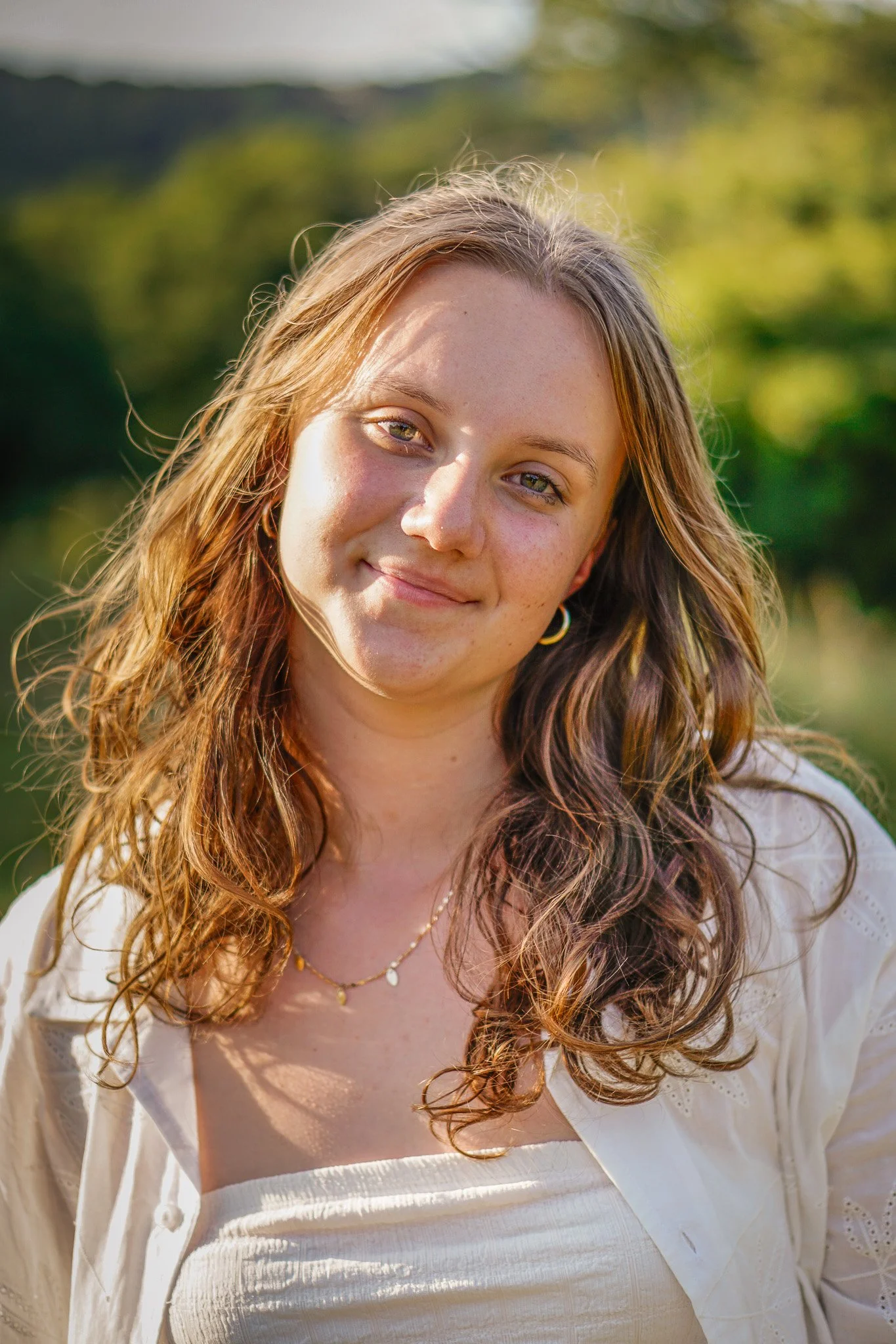 A young woman with long, wavy brown hair and light skin, smiling softly outdoors in natural light, with a blurred green background.