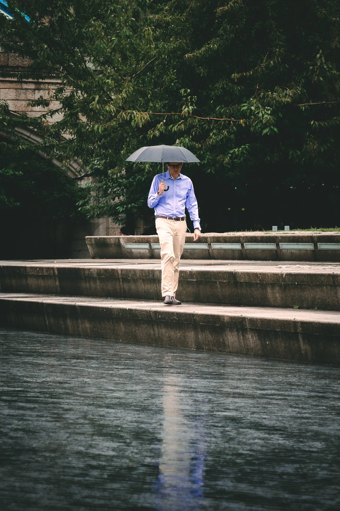 A man in a blue shirt and beige pants walking down steps by water, holding an umbrella during a rainy day.