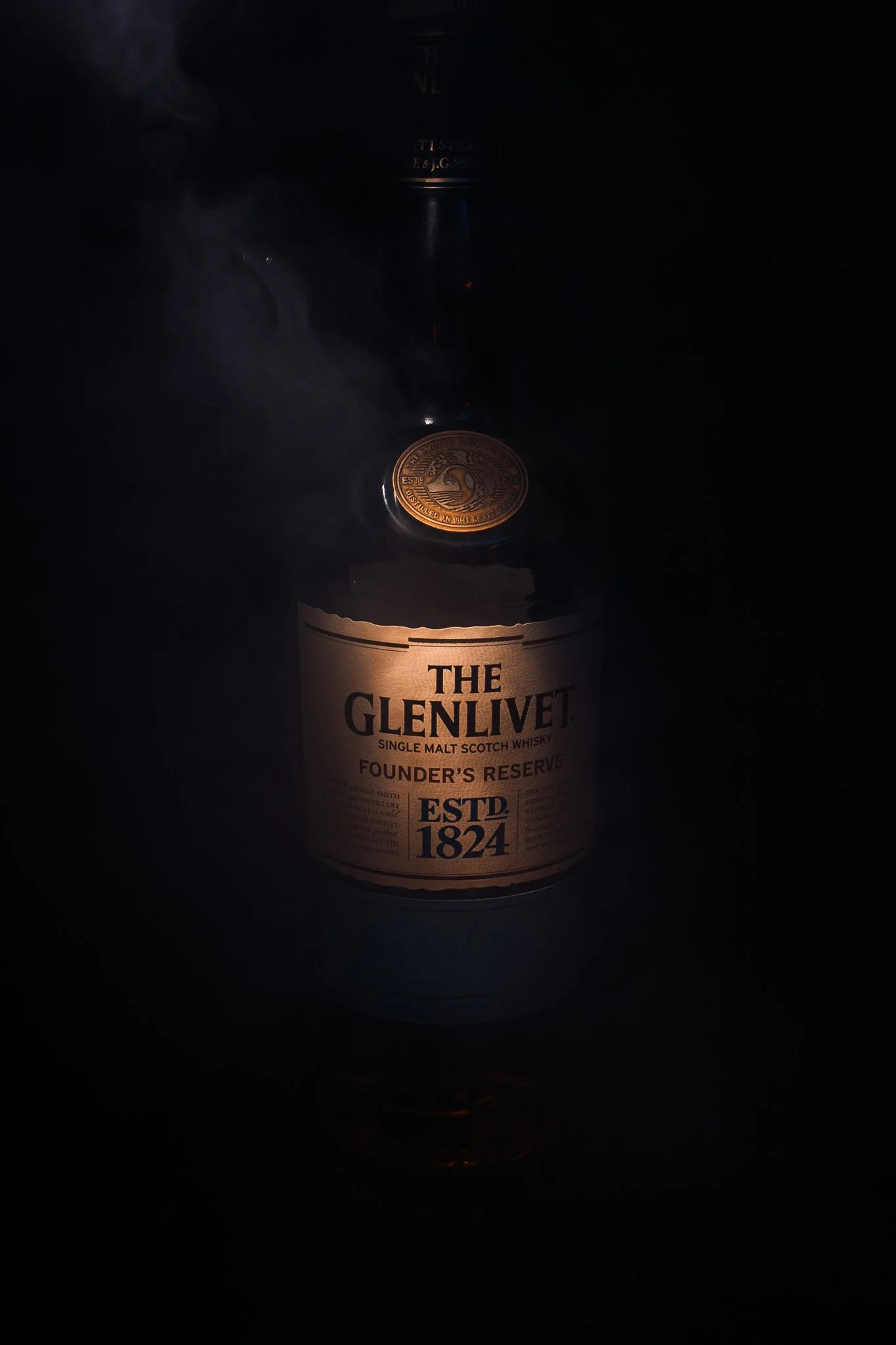 A bottle of The Glenlivet Single Malt Scotch Whisky labeled 'Founder's Reserve' with a gold seal on the neck, illuminated slightly in dark lighting with some smoke or vapor around it.