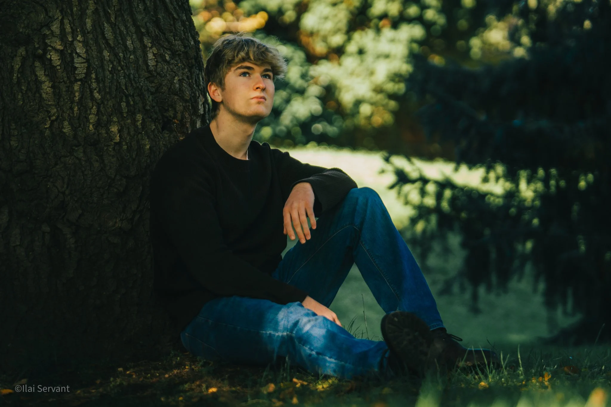 A young man sitting on the grass and leaning against a tree, gazing thoughtfully into the distance, in a lush outdoor setting with trees and greenery in the background.