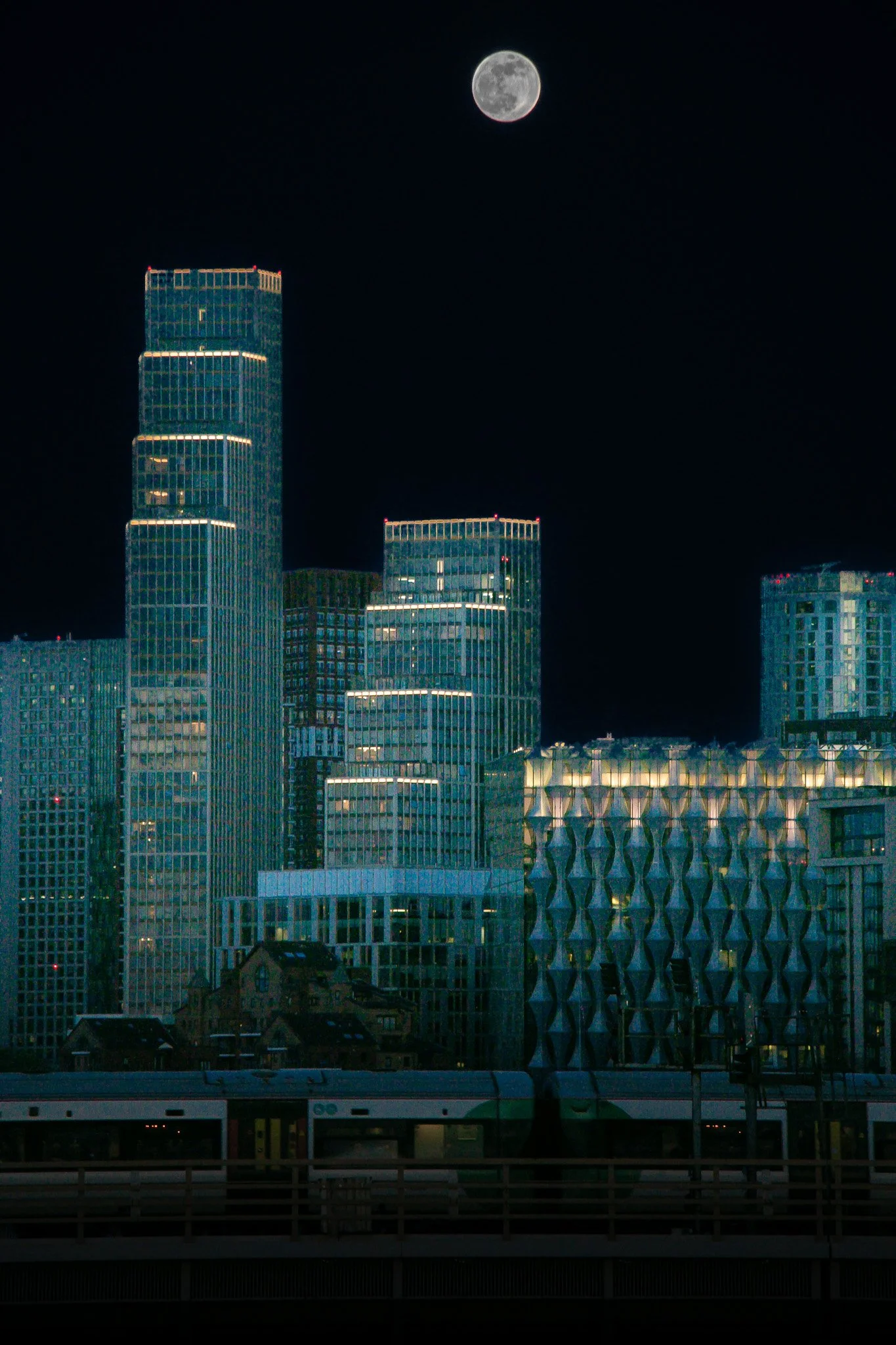 Nighttime cityscape with illuminated skyscrapers and a full moon in the clear sky.