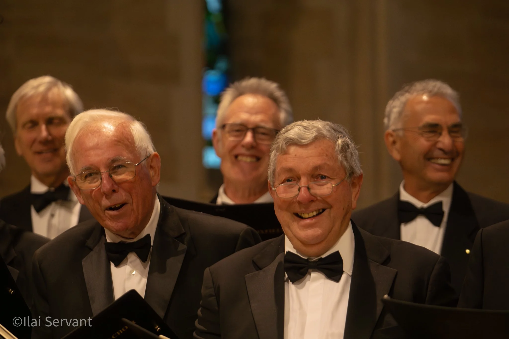 A group of elderly men in tuxedos, some wearing glasses, smiling and singing in a choir or an orchestra, in a dimly lit church or concert hall.