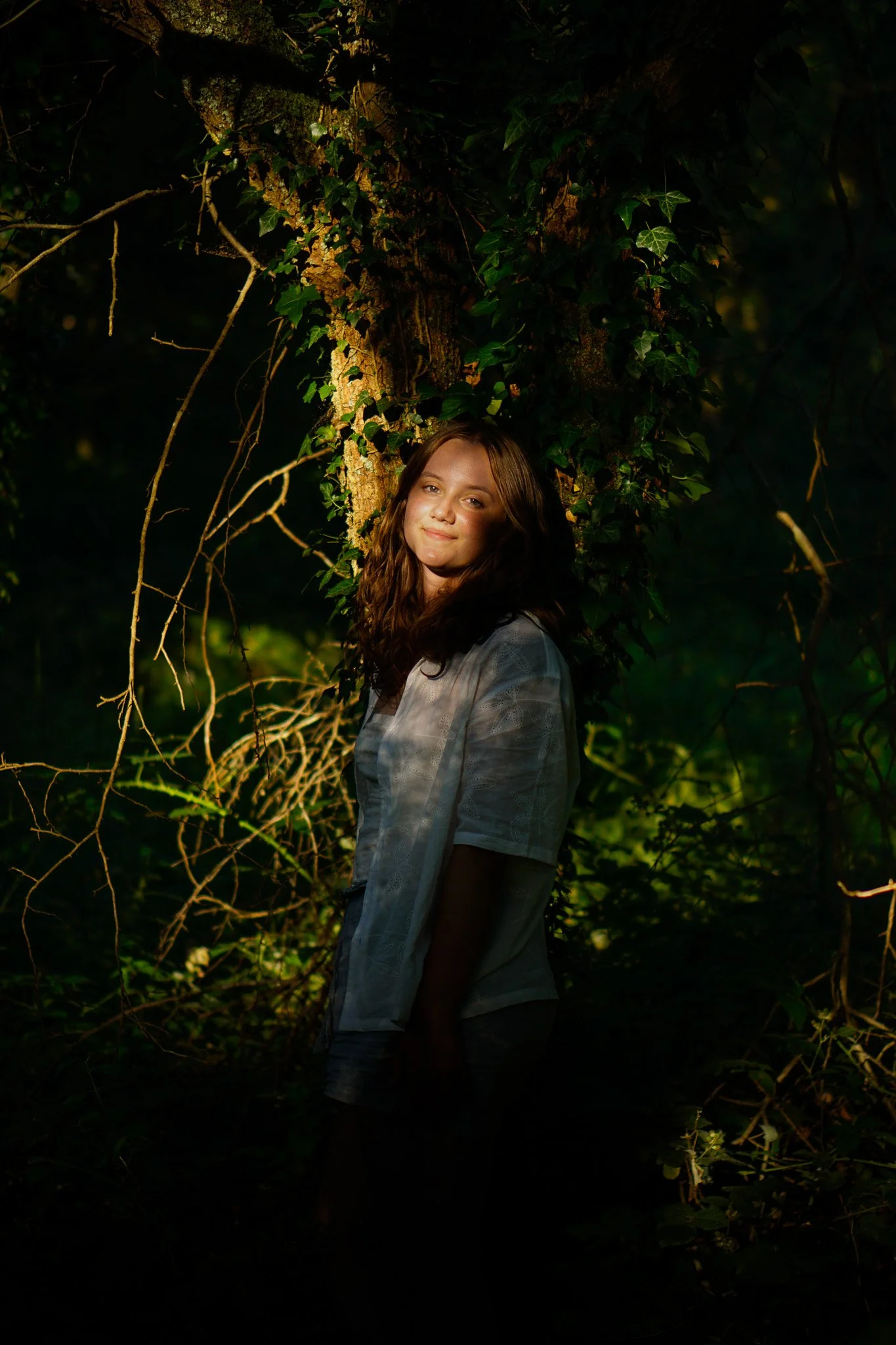 A young woman with wavy brown hair smiling softly, standing outdoors in a forested area at night, illuminated by a warm light.