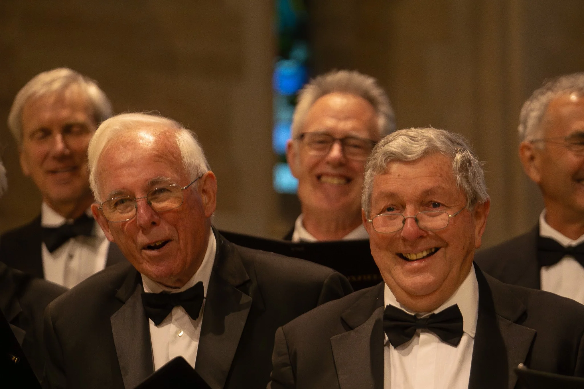 Group of elderly men dressed in formal tuxedos and bow ties, smiling and performing in a choir or musical ensemble inside a church or venue with stained glass window.