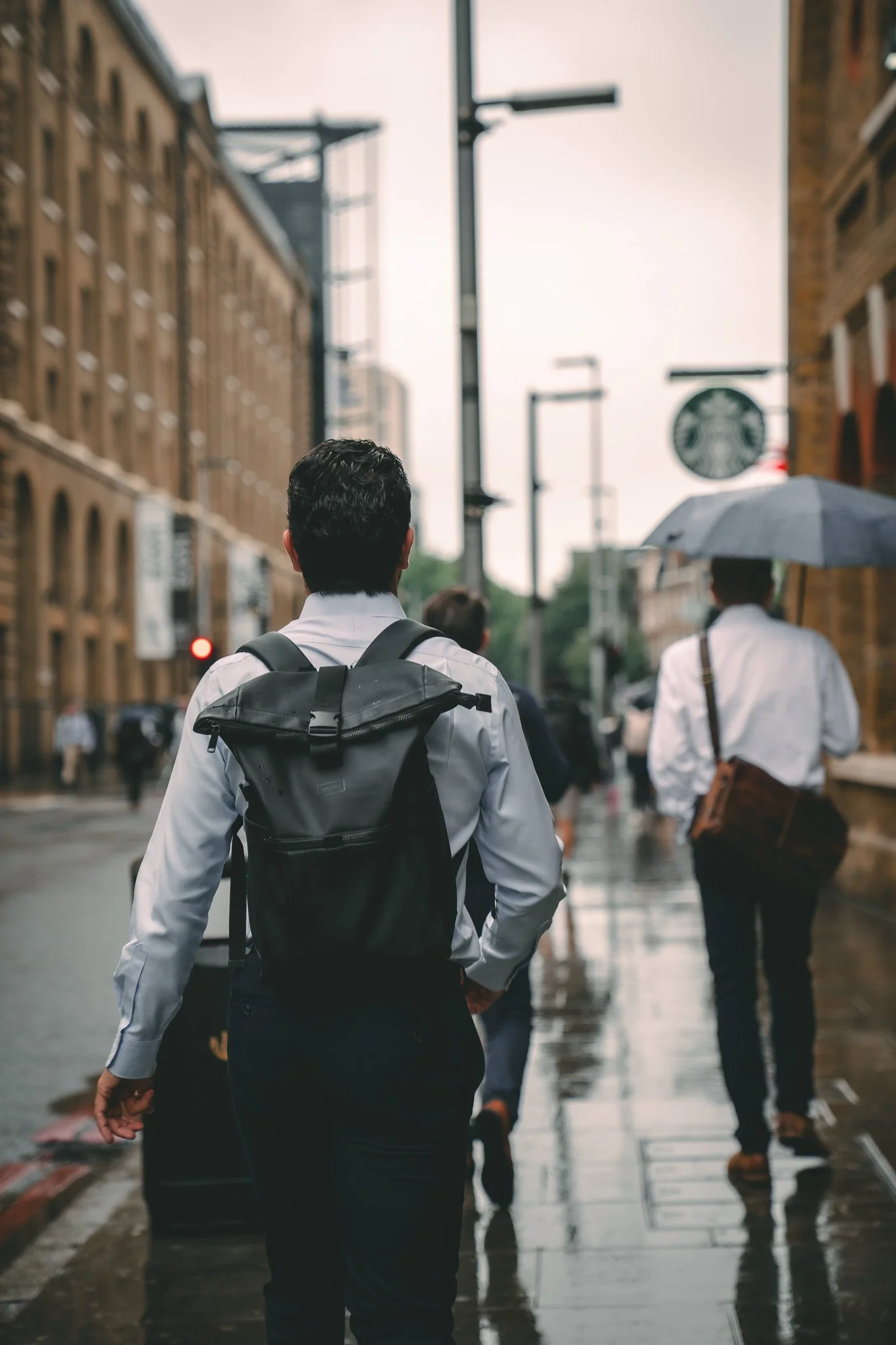 People walking on a rainy city street, some with umbrellas, wet pavement, city buildings, and streetlights.