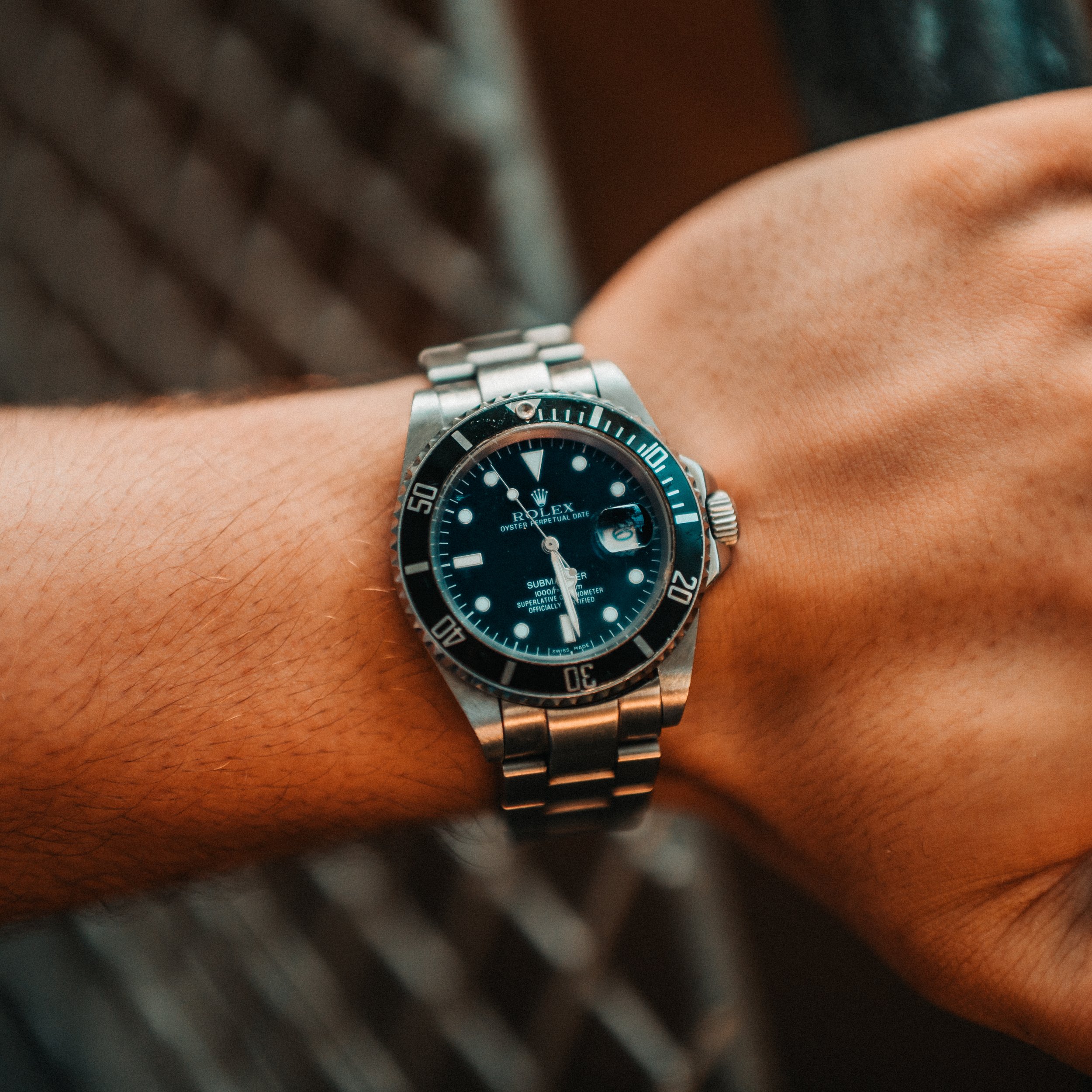 Close-up image of a person's wrist wearing a silver Rolex watch with a black dial and rotating bezel, showing the time and date.