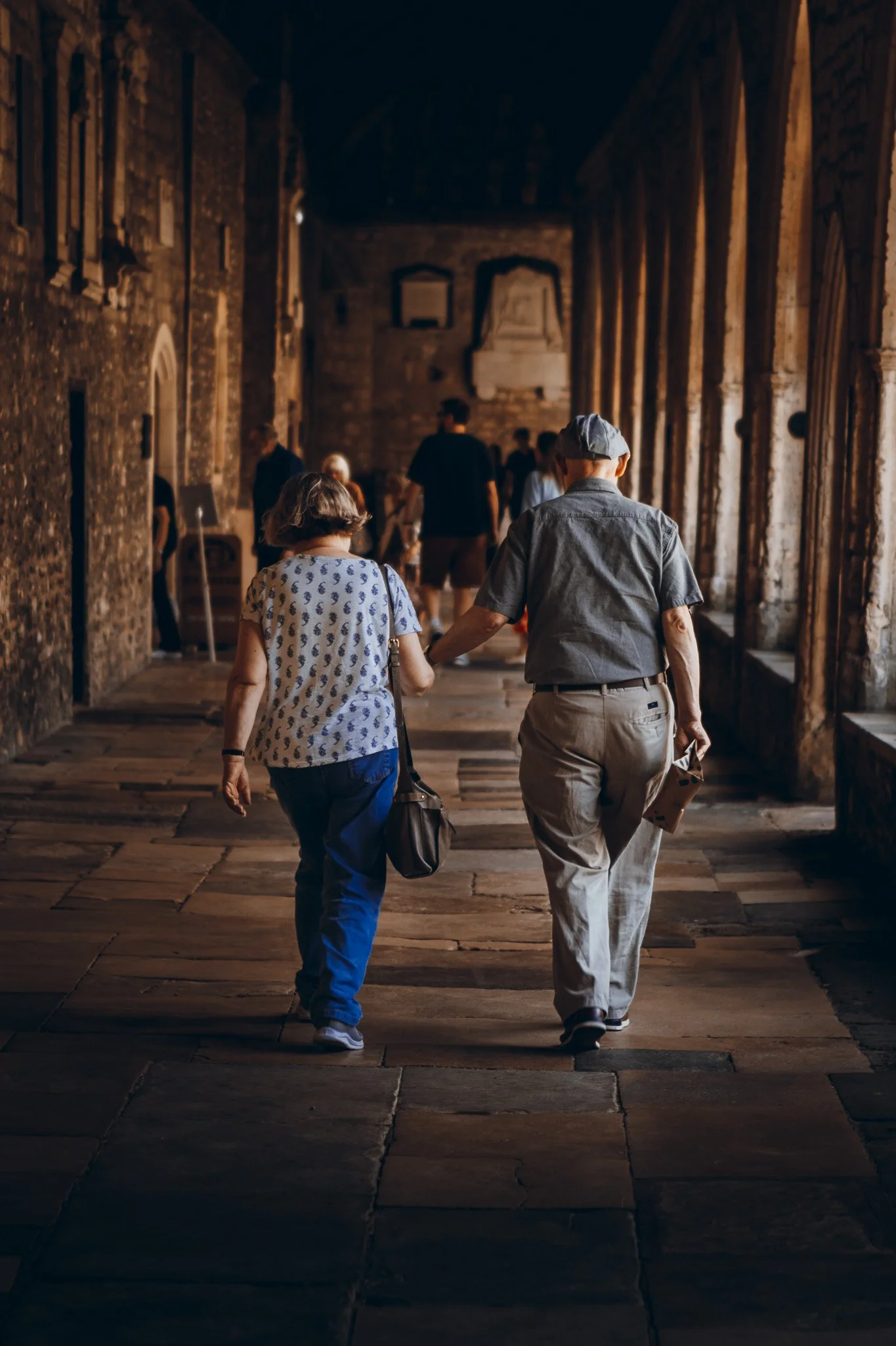 A senior couple walking hand in hand through an old stone corridor with large arched windows and several other visitors in the background.